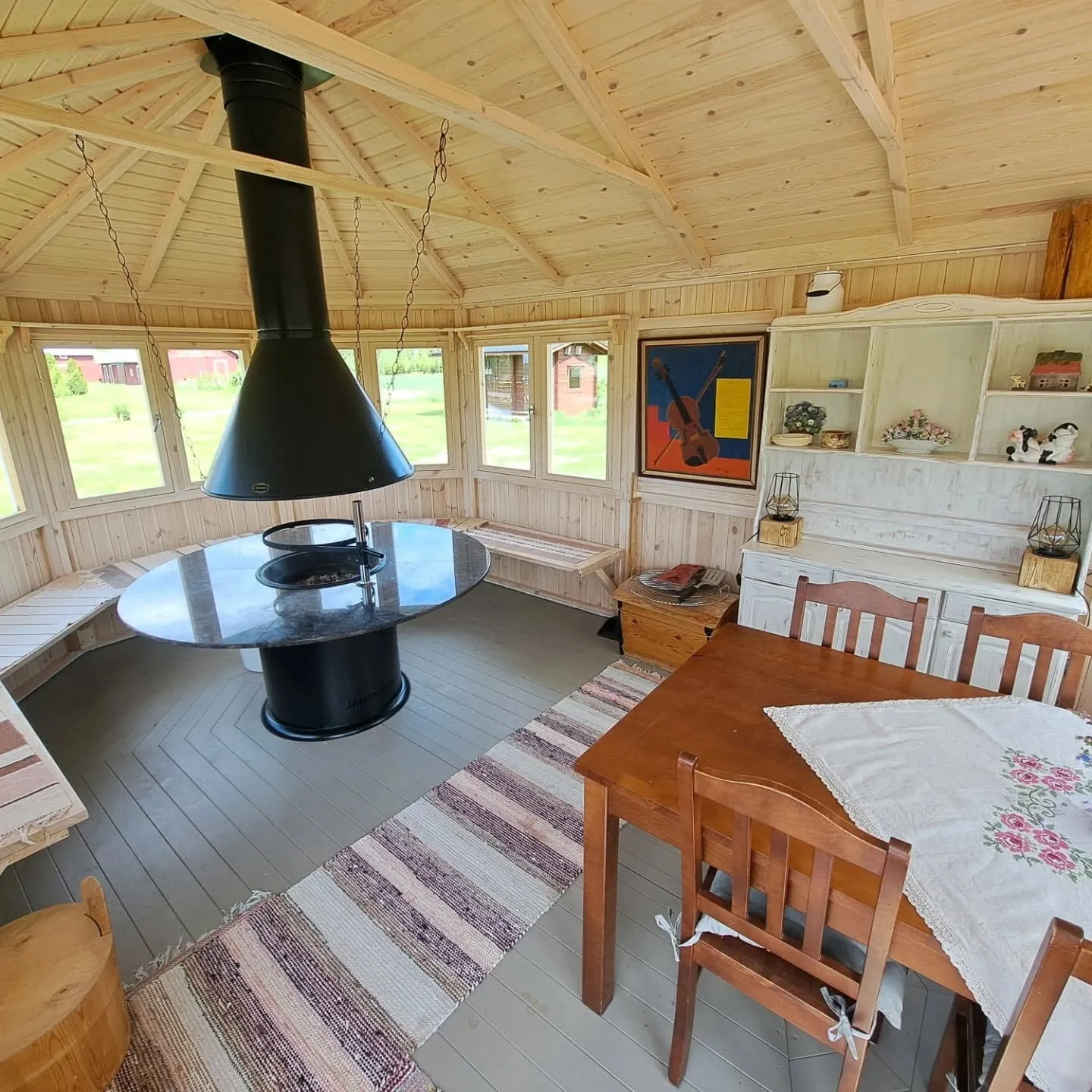 Valoisa puinen grillikota, jossa on vaaleat puuseinät ja katto. Keskellä on musta savupiippu ja sen alla pyöreä pöytä sisäänrakennetulla grillillä. Pöydän ympärillä on puupenkit. Oikealla on ruskea ruokapöytä neljällä tuolilla, pöydällä valkoinen kukkakuvioinen pöytäliina. Seinällä on viuluja esittävä taulu ja valkoinen hyllykkö esineineen. Harmaalla puulattialla on raidallinen matto. Ikkunoista näkyy vihreää nurmikkoa.