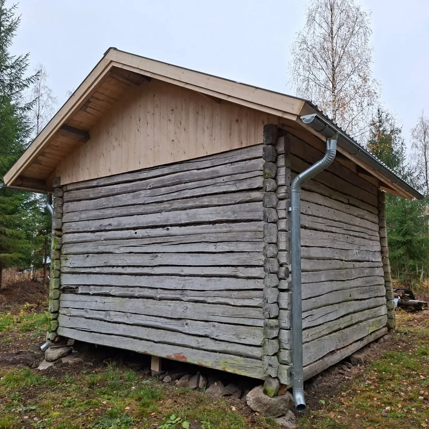 Vanha, harmaa hirsirakennus, jossa on uusi vaalea puinen päätykatto ja hopeanvärinen sadevesijärjestelmä. Rakennuksen seinät ovat sään kuluttamia, vaakasuoria hirsiä. Se seisoo kivien ja puupalojen varassa. Maassa on ruohoa ja lehtiä, ja taustalla näkyy puita pilvisen taivaan alla.