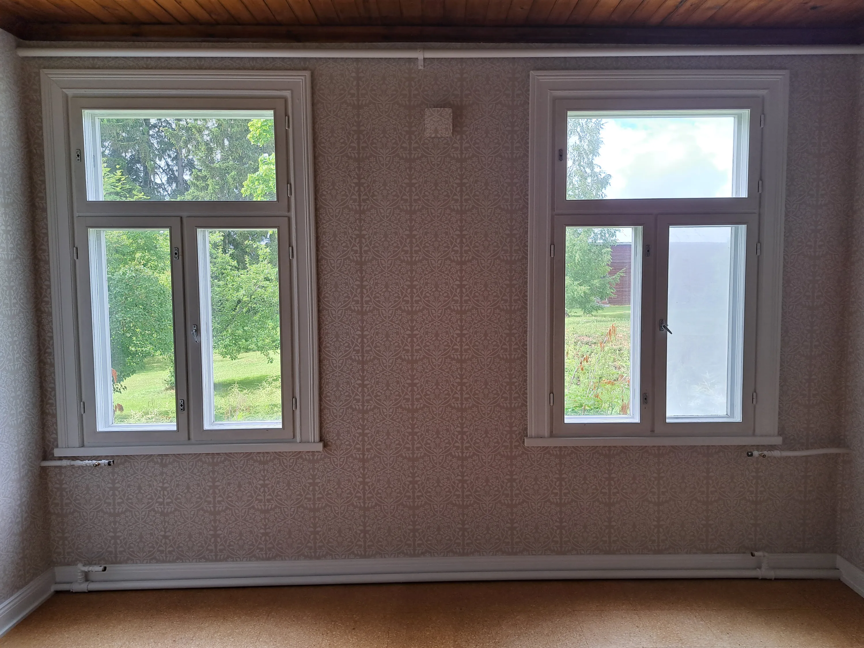 Tyhjä huone, jossa on kaksi valkoista ikkunaa. Seinillä on vaaleanruskea tapetti toistuvalla kasviaiheisella kuviolla. Kattona on tumma puupaneeli ja lattiana vaaleanruskea korkkilattia. Valkoiset putket kulkevat seinillä ylhäällä ja alhaalla. Ikkunoista näkyy vihreää puustoa ja nurmikkoa; oikeanpuoleisen ikkunan alalasi on osittain peitetty. Huoneessa on kirkas luonnonvalo.