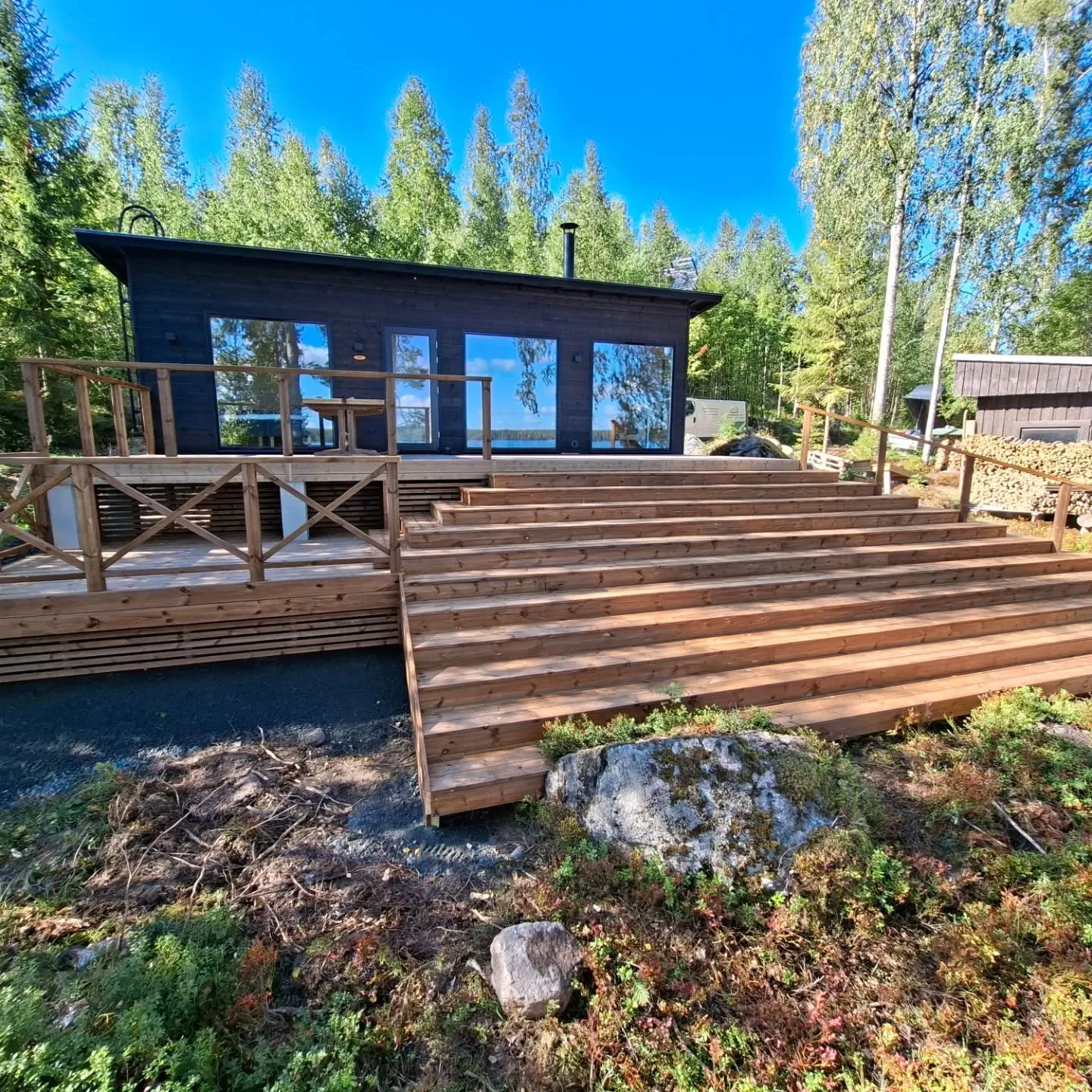 Aurinkoisena päivänä kuvattu tumma puurakennus, jossa on suuret ikkunat heijastaen sinistä taivasta ja järvimaisemaa. Rakennuksen edessä on puinen terassi kaiteilla ja leveät puuportaat, jotka laskeutuvat alas maastoon. Ympärillä on tiheä metsä ja oikealla näkyy pienempi puinen varasto halkopinon kera. Maasto on kivinen ja siinä kasvaa matalaa kasvillisuutta.