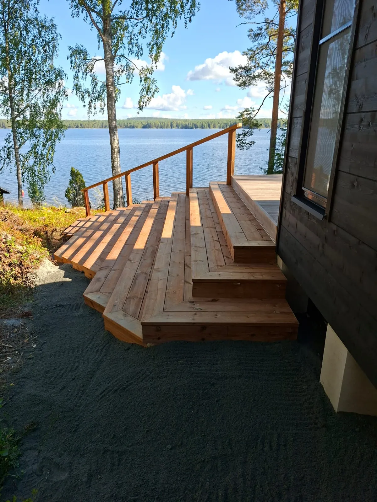 Aurinkoinen kuva puuterassista ja portaista, jotka laskeutuvat kohti järveä. Terassi on vaaleaa puuta ja siinä on puinen kaide. Oikealla on tummanruskea talon seinä ikkunalla. Taustalla avautuu sininen järvimaisema metsäisine rantoineen ja vaaleansininen taivas valkoisine pilvineen. Terassin ympärillä maassa on tummaa sepeliä. Auringonvalon luomia varjoja näkyy terassin laudoituksella.