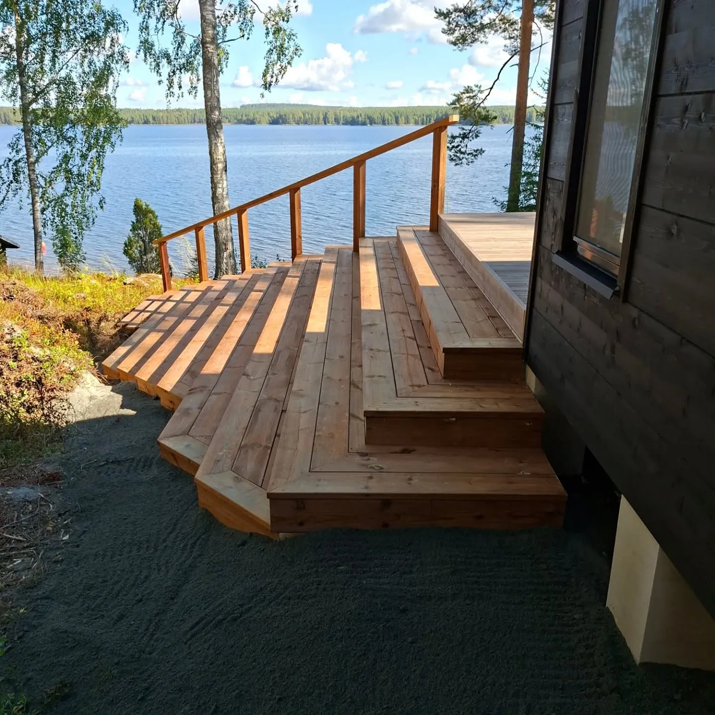 Aurinkoinen kuva vaaleanruskeasta puuterassista ja portaista, jotka laskeutuvat tumman rakennuksen vierestä kohti järveä. Terassilla on puinen kaide. Vasemmalla näkyy vihreää lehdistöä. Taustalla on laaja sininen järvi metsäisellä rannalla ja yläpuolella sininen taivas valkoisine pilvineen. Maassa terassin edessä on tummaa mursketta. Auringonvalo luo pitkiä varjoja terassin pinnalle.