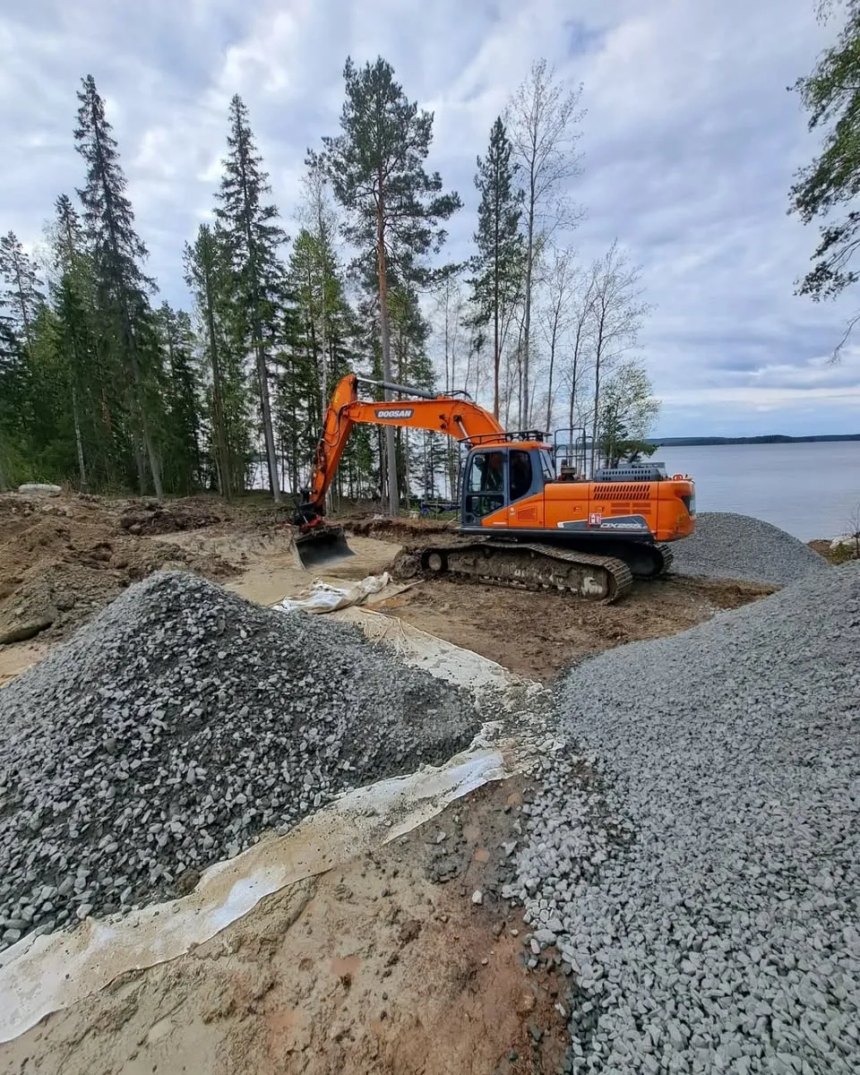 Kuvassa näkyy oranssi Doosan DX225LC -kaivinkone rakennustyömaalla järven rannalla pilvisenä päivänä. Etualalla on useita kasoja harmaata mursketta ja ruskeaa maata, joiden seassa on valkoista suodatinkangasta. Kaivinkone on kuvan keskellä oikealla, sen kauha on laskettuna maahan. Taustalla vasemmalla kohoaa tiheä metsä korkeita mäntyjä ja muita puita. Oikealla taustalla avautuu laaja järvimaisema pilvisen, harmaan taivaan alla.