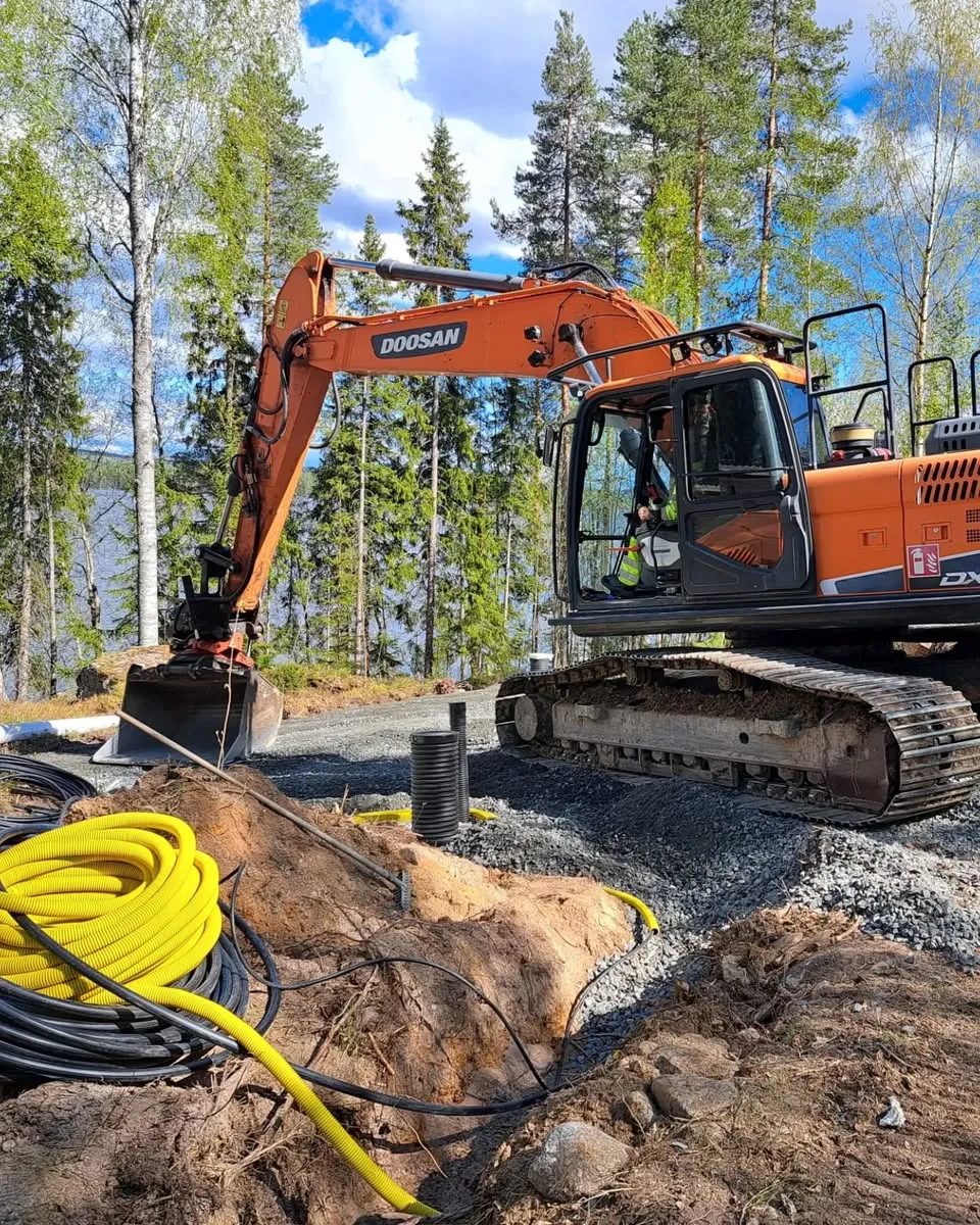 Oranssi Doosan-kaivinkone seisoo soraisella maalla aurinkoisena päivänä. Sen edessä on kaivettu oja, jossa kulkee keltaisia putkia ja mustia kaapeleita. Taustalla näkyy metsää ja järvi sinisen, pilvisen taivaan alla.