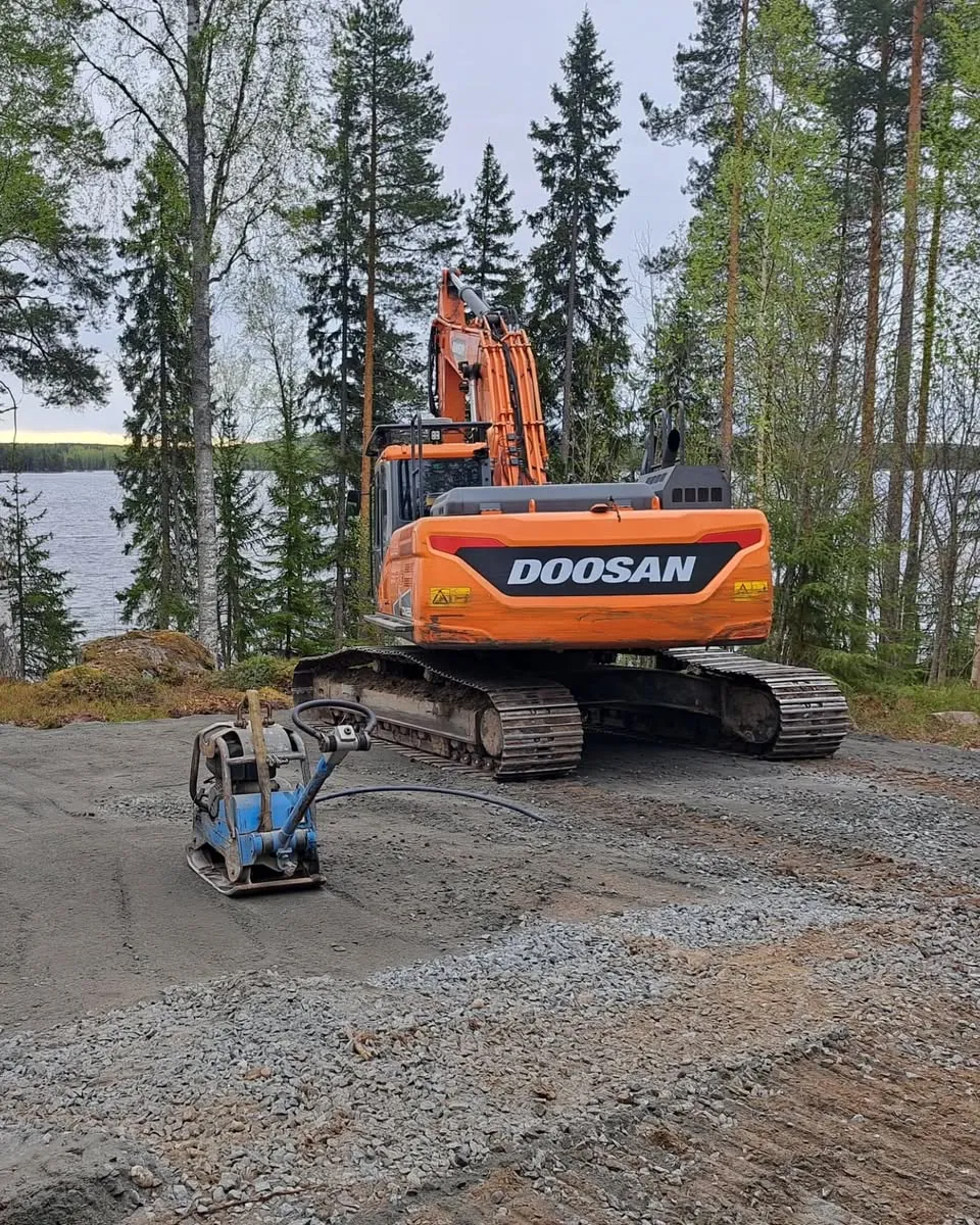 Oranssi Doosan-kaivinkone seisoo soraisella tiellä, jonka edustalla on sininen tärytin tiivistämässä maata. Taustalla on järvi ja tiheä metsä pilvisen taivaan alla.