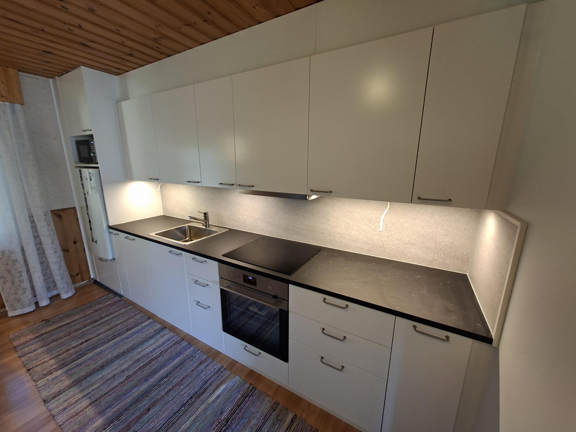 Moderni keittiö, jossa on valkoiset ylä- ja alakaapit sekä tummanharmaa työtaso. Vasemmalla on tiskiallas ja oikealla induktioliesi integroidun uunin yläpuolella. Kaappien alusvalaistus valaisee vaaleanharmaata välitilaa. Vasemmalla näkyy mikroaaltouuni jääkaapin päällä. Katossa on puupaneelit ja lattialla raidallinen matto puulattialla.