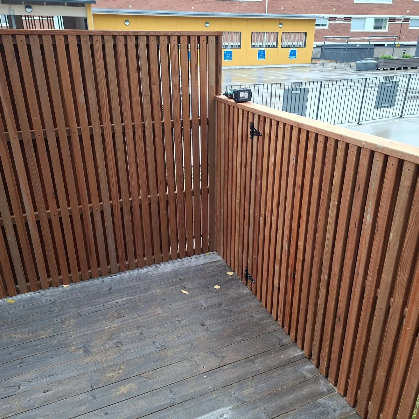 Kuva esittää puista kulma-aitaa ja tummaa puuterassia. Aita on tehty pystysuorista, punertavanruskeista laudoista. Terassin lattia on tummanharmaata puuta, ja siinä on muutama pudonnut lehti. Oikeanpuoleisen aidan yläreunalla on musta akkuporakone. Aidassa on myös mustia metallikiinnikkeitä. Aidasta ulospäin näkyy asfaltoitu piha-alue, metalliaitoja sekä keltaisia ja tiilisiä rakennuksia taustalla. Valaistus on tasaista päivänvaloa.