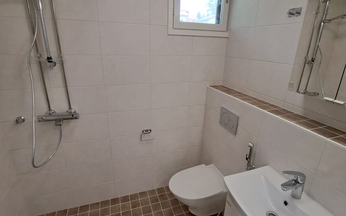 Valoisa kylpyhuone, jossa on vaaleat seinälaatat ja tummanruskeat lattialaatat. Vasemmalla on krominen suihku. Keskellä on valkoinen seinä-WC, jonka yläpuolella on ruskeilla laatoilla päällystetty taso ja huuhtelupainike. Oikealla on valkoinen pesuallas kromisella hanalla ja bideesuihkulla, sekä peili. Ylhäällä keskellä on valkoinen ikkuna.