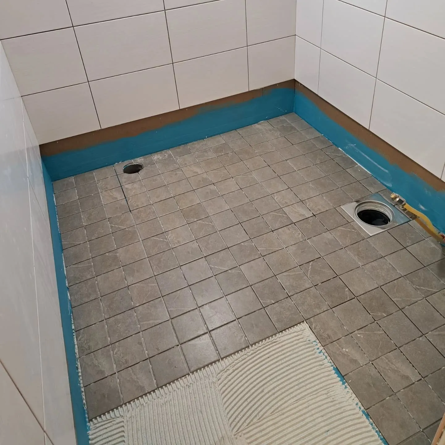 Kylpyhuoneen lattiaa laatoitetaan. Valkoiset seinälaatat on asennettu. Lattialla on harmaita neliönmuotoisia laattoja, ja osassa lattiaa näkyy valkoista laattaliimaa. Seinien alaosassa ja lattian reunoilla on sininen vedeneristyskerros. Lattiassa on kaksi viemäriaukkoa, joista toisessa on metallinen lattiakaivo ja keltainen letku.