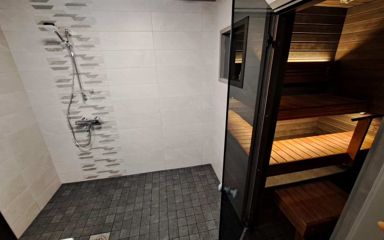 Kylpyhuone, jossa on suihku ja sauna. Vasemmalla on suihkualue vaaleanharmailla seinälaatoilla, joissa on pystysuora kuvioitu laattaraita. Lattiassa on tummanharmaat vinoneliön muotoiset laatat. Oikealla on tummakehyksinen lasiovi, joka avautuu saunaan. Saunan sisällä on puuseinät ja -penkit lämpimällä, epäsuoralla valaistuksella. Suihkun ja saunan välissä on pieni ikkuna.