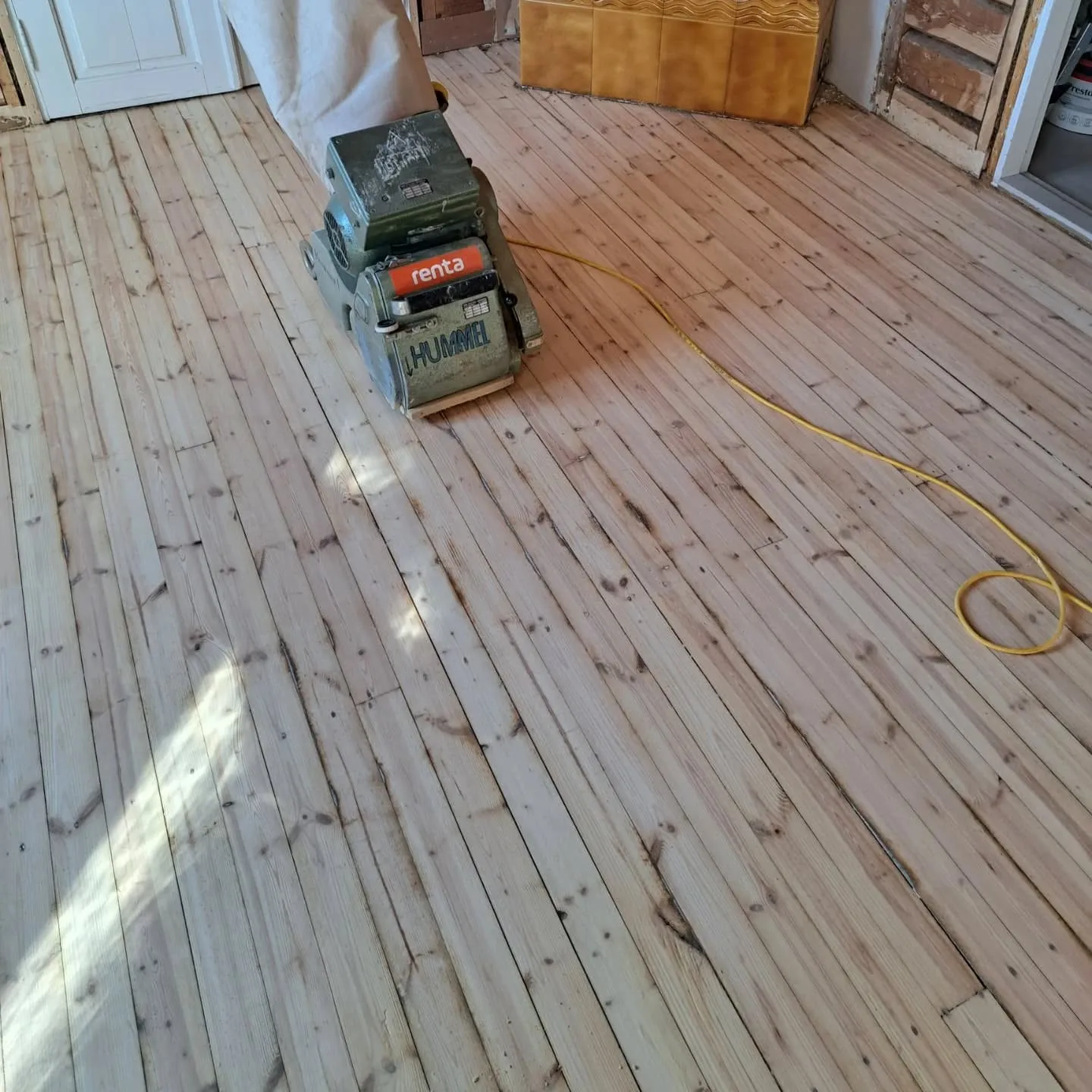 Kuva esittää lattianhiontakonetta, joka seisoo puulattialla. Lattia on osittain hiottu: vasemmalla on vaaleaa, hiottua puuta ja oikealla tummempaa, hiomatonta puuta. Oliivinvihreässä koneessa lukee "HUMMEL" ja "renta". Keltainen sähköjohto kulkee lattialla. Taustalla näkyy valkoinen ovi, laatoitettu seinä ja puurunko. Luonnonvalo valaisee lattiaa vasemmalta.