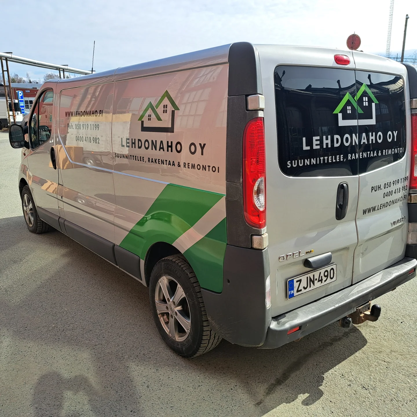 Hopeanvärinen Opel Vivaro -pakettiauto on pysäköity aurinkoisena päivänä. Auton kyljessä ja takaikkunassa on vihreä-musta Lehdonaaho Oy:n logo, jossa on kaksi taloa, sekä teksti 'SUUNNITTELEE, RAKENTAA & REMONTOI'. Yhteystiedot, kuten puhelinnumerot ja verkkosivusto, näkyvät myös. Maassa näkyy auton varjoja.