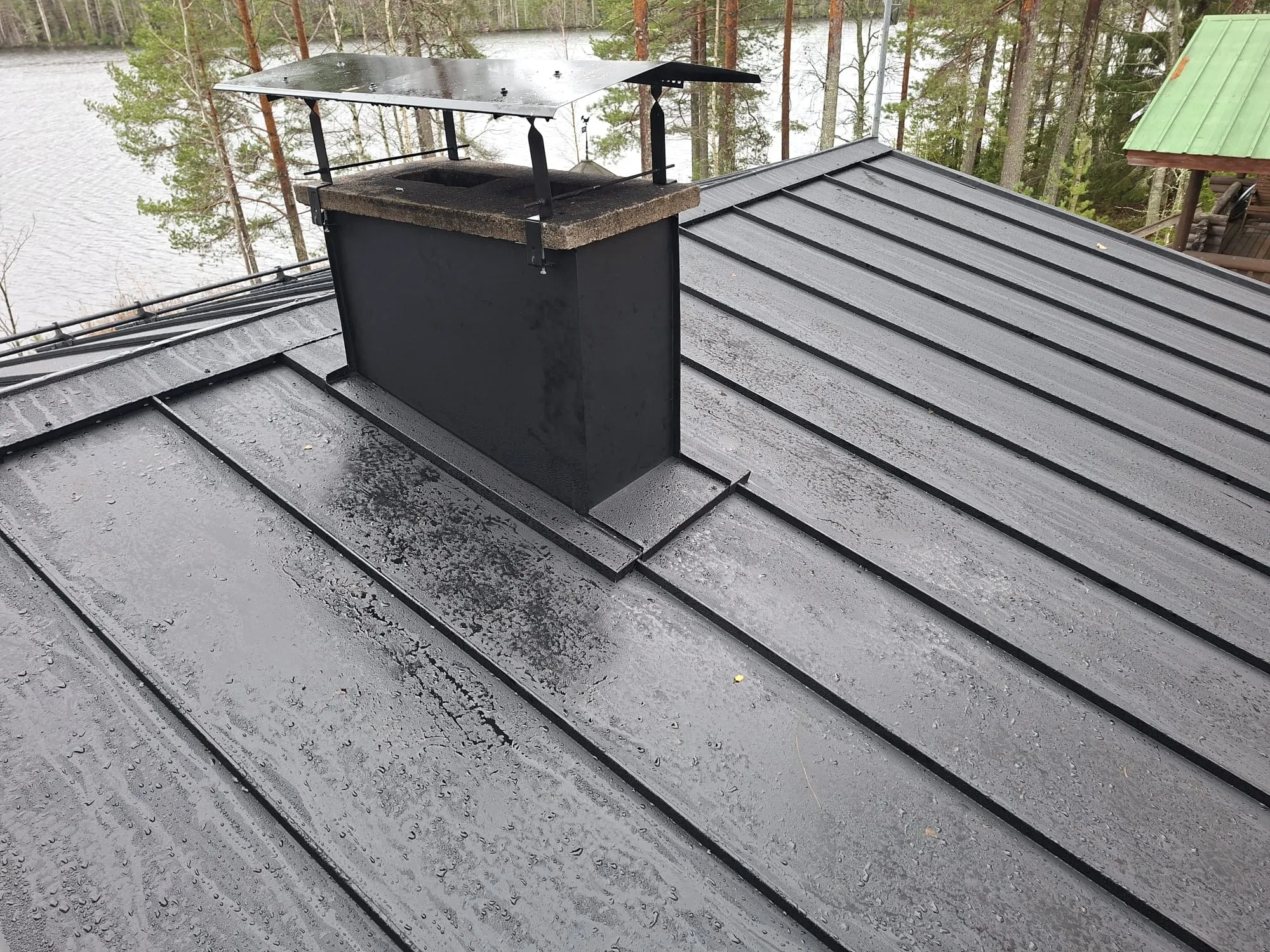 Kuvassa näkyy märkä, musta peltikatto, jossa on runsaasti vesipisaroita. Katon keskellä on musta savupiippu, jonka päällä on kivinen yläosa ja metallinen sadehattu. Taustalla avautuu järvimaisema metsineen. Oikealla näkyy osittain vihreäkattoinen hirsinen rakennus.