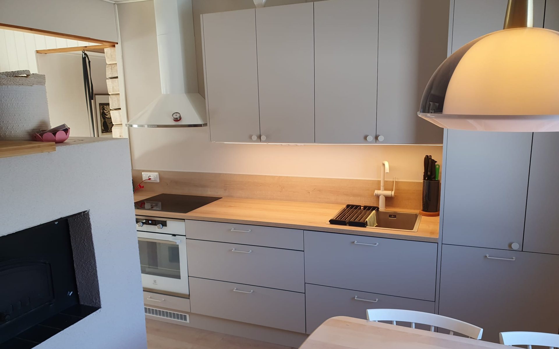 Moderni keittiö ja ruokailutila, jossa on vaaleanharmaat kaapit, puunvärinen työtaso ja valkoinen liesituuletin. Työtason yläpuolella on lämmin valaistus. Kuvassa näkyy myös valkoinen takka, jossa on musta aukko, sekä osittain puinen ruokapöytä ja valkoiset tuolit. Oikealla roikkuu suuri, vaalea kattolamppu.