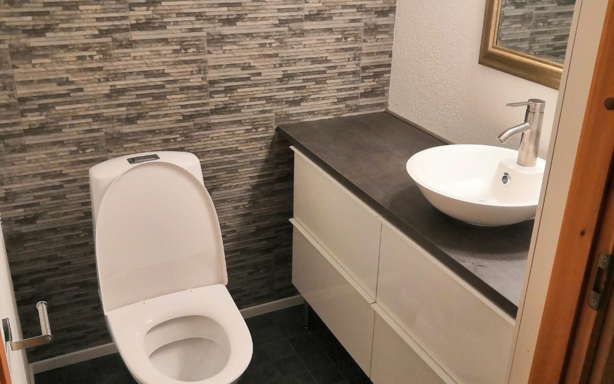 Moderni WC-tila, jossa vasemmalla on valkoinen WC-istuin kansi auki. Sen takana on seinä, jossa on tummanharmaita ja ruskeita kivimäisiä laattoja. Oikealla on tummanharmaa työtaso, jonka alla on valkoinen allaskaappi kahdella laatikolla. Työtasolla on valkoinen pyöreä pesuallas ja hopeinen hana. Oikealla ylhäällä näkyy osittain kultakehyksinen peili. Lattia on tummanharmaata laattaa.