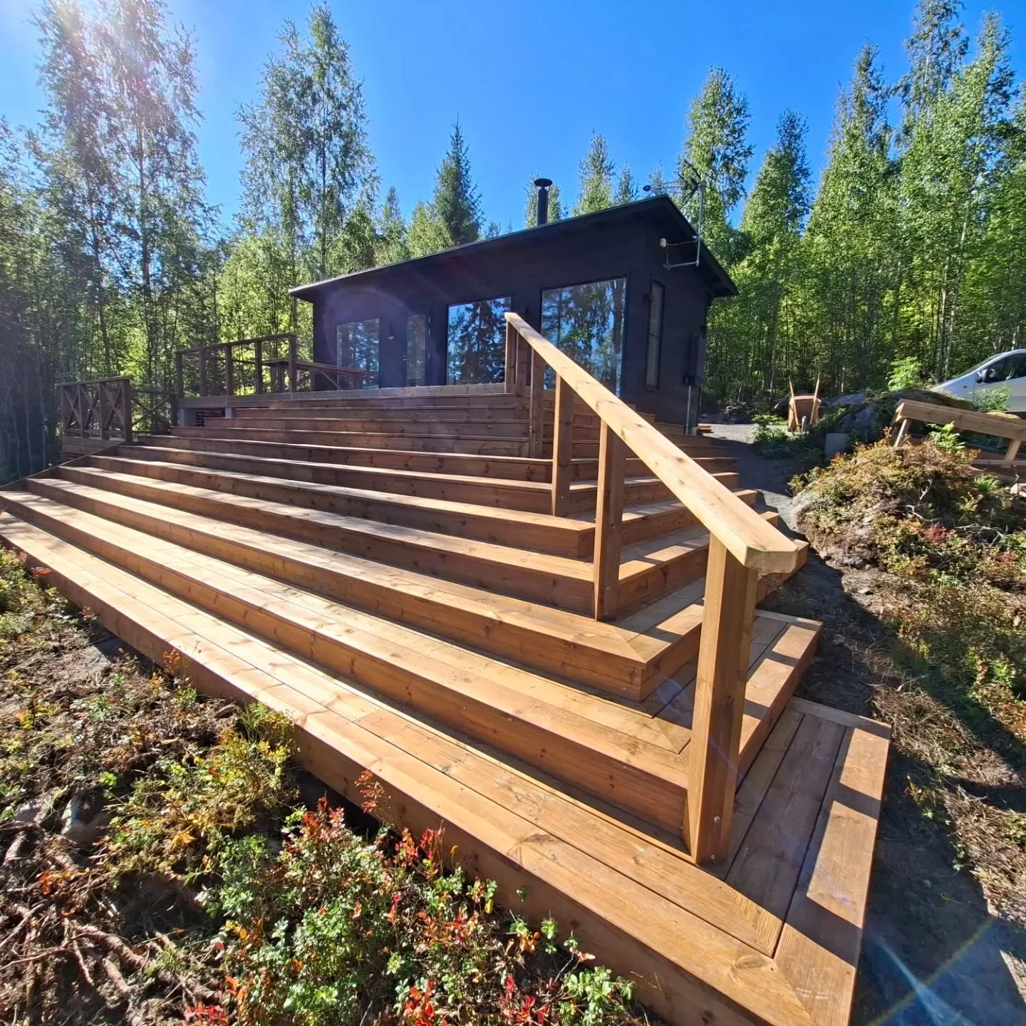 Aurinkoisena päivänä tummanruskea mökki seisoo metsän keskellä. Mökille johtaa laaja, useita tasoja käsittävä puinen portaikko ja terassi, jossa on puukaiteet. Kirkkaan sininen taivas ja puiden läpi siivilöityvä auringonvalo valaisevat maisemaa. Portaikon vieressä kasvaa vihreää ja punertavaa kasvillisuutta. Oikealla taustalla näkyy osittain valkoinen auto ja kottikärryt.
