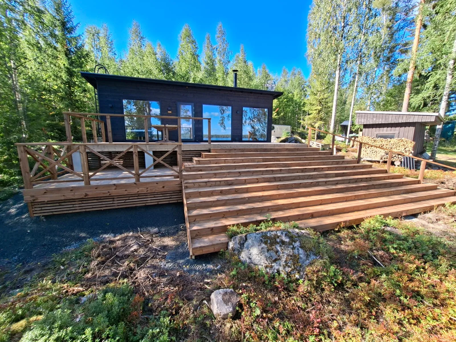 Moderni tumma mökki metsäisessä maisemassa, jossa on suuret ikkunat heijastaen sinistä taivasta ja vettä. Edessä on laaja puinen terassi kaiteineen ja leveät puuportaat. Oikealla takana näkyy pieni puuvaja halkopinoineen. Ympärillä on korkeita puita, aluskasvillisuutta ja kiviä kirkkaassa auringonvalossa.