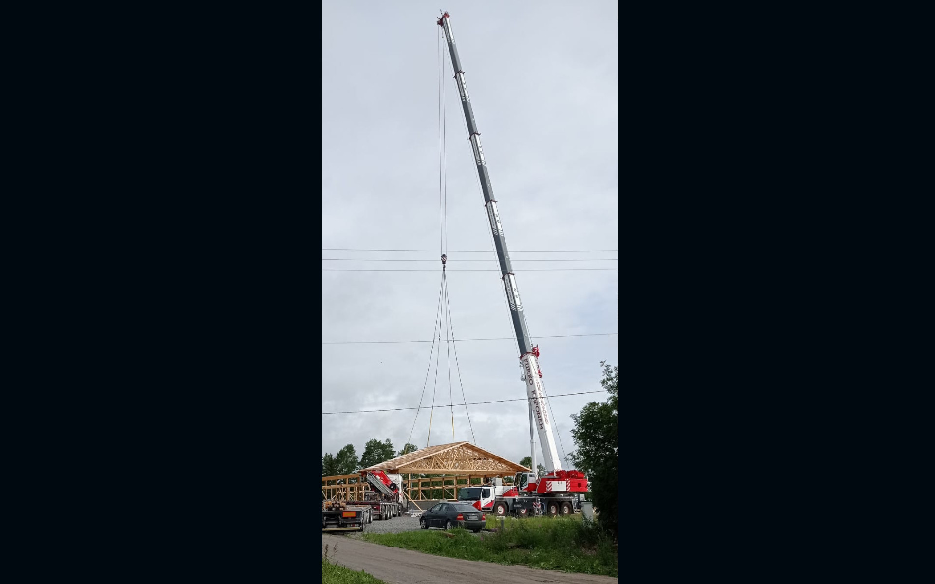Suuri punavalkoinen ajoneuvonosturi, jonka puomissa lukee "PURO KROPONEN", nostaa puista kattoristikkoa rakenteilla olevan puurakennuksen rungon päälle. Kuvan etualalla on hiekkatie ja vihreää ruohoa. Taustalla näkyy sähkölinjoja, puita ja pilvinen taivas. Paikalla on myös muita rakennustyömaan ajoneuvoja ja tumma henkilöauto.