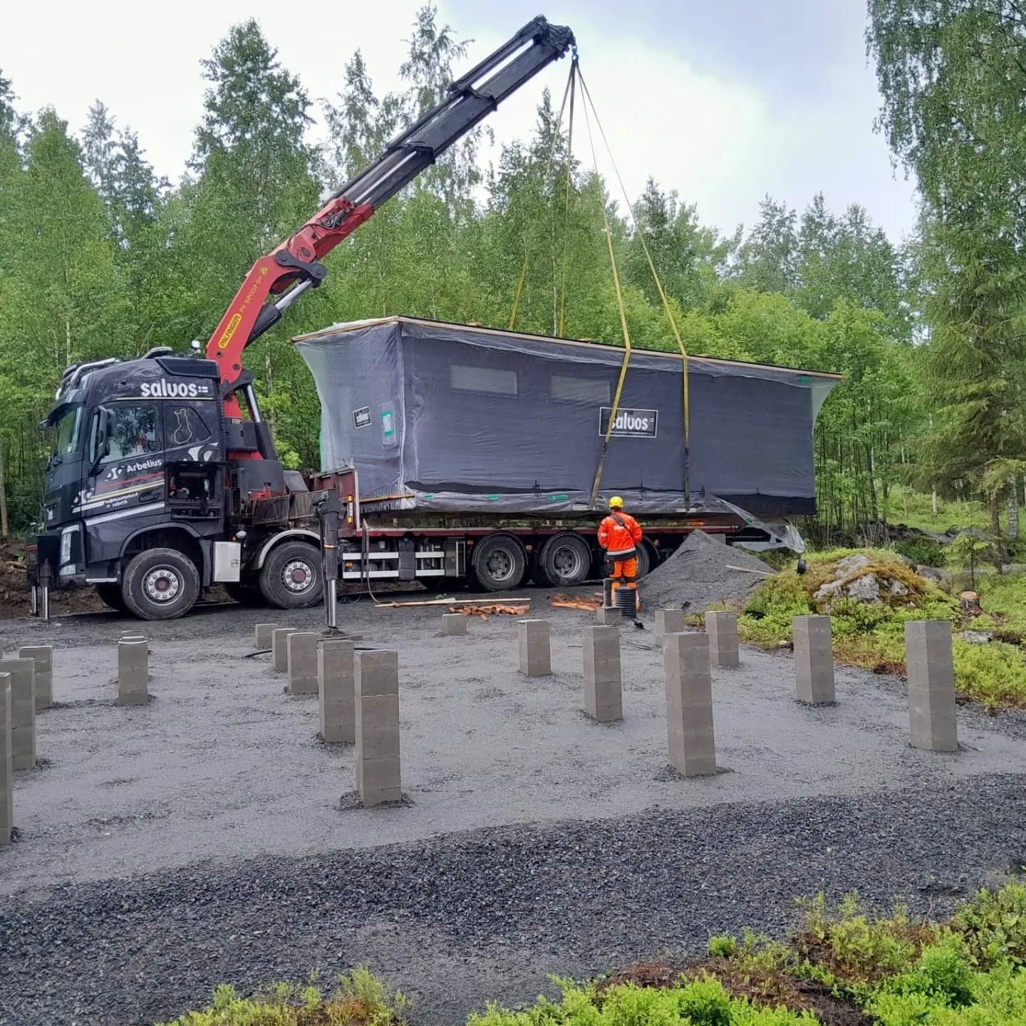Kuorma-auto nosturilla nostaa tummanharmaata talomoduulia rakennustyömaalla. Moduuli on kääritty suojamateriaaliin ja siinä on "saluos"-logo. Oranssipukuinen työntekijä keltaisessa kypärässä ohjaa moduulia. Maassa on harmaita betonipilareita sorapedillä. Taustalla on vihreää metsää ja pilvinen taivas.