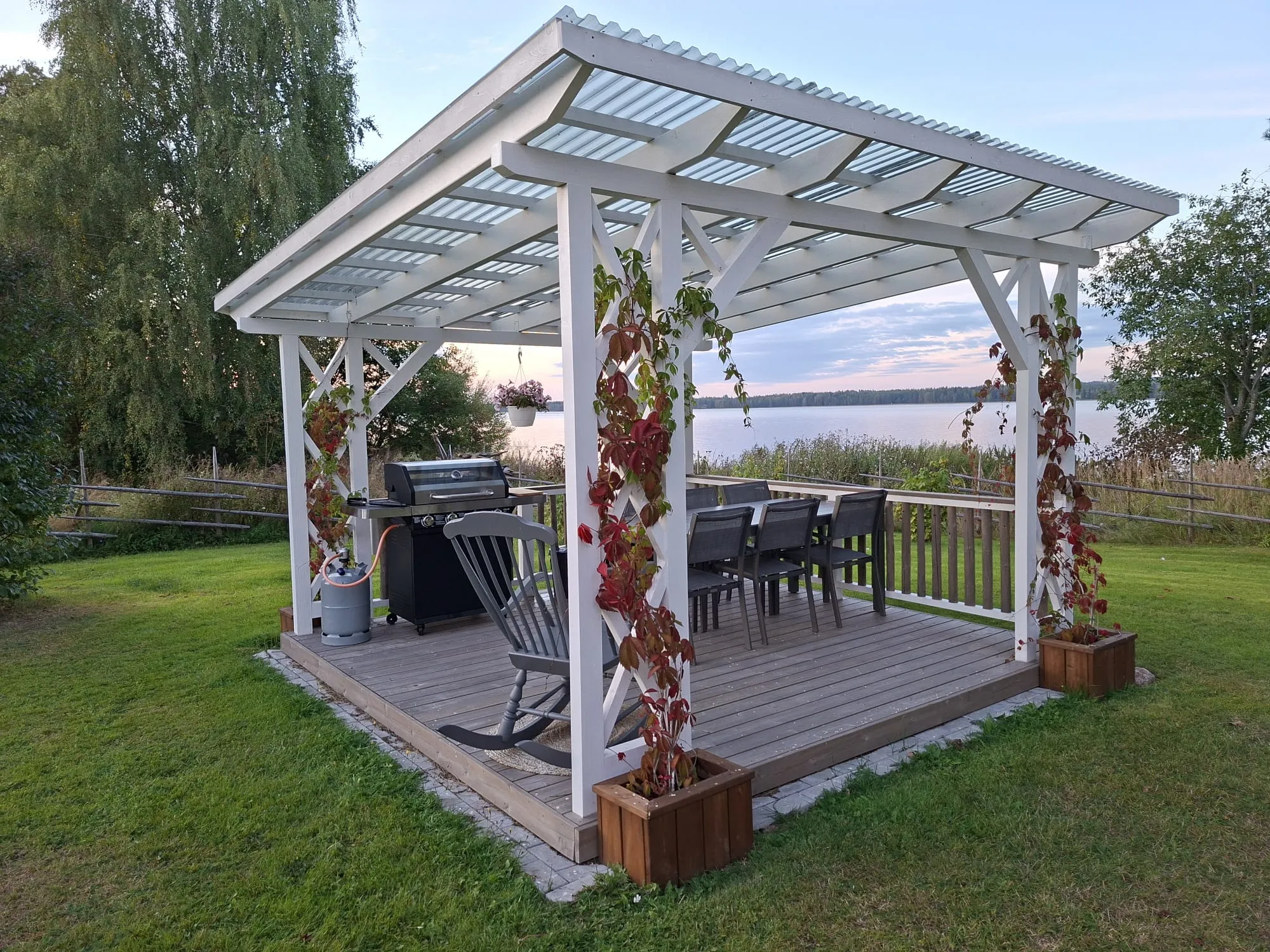 Valkoinen puinen pergola läpinäkyvällä aaltokatolla seisoo nurmikolla järven rannalla. Sen sisällä on puinen terassi, kaasugrilli, keinutuoli ja ruokapöytä tuoleineen. Punalehtiset ja vihreät köynnökset kasvavat pylväitä pitkin, ja katosta roikkuu kukkaruukku. Taustalla näkyy järvi ja puita ilta-auringon valossa.