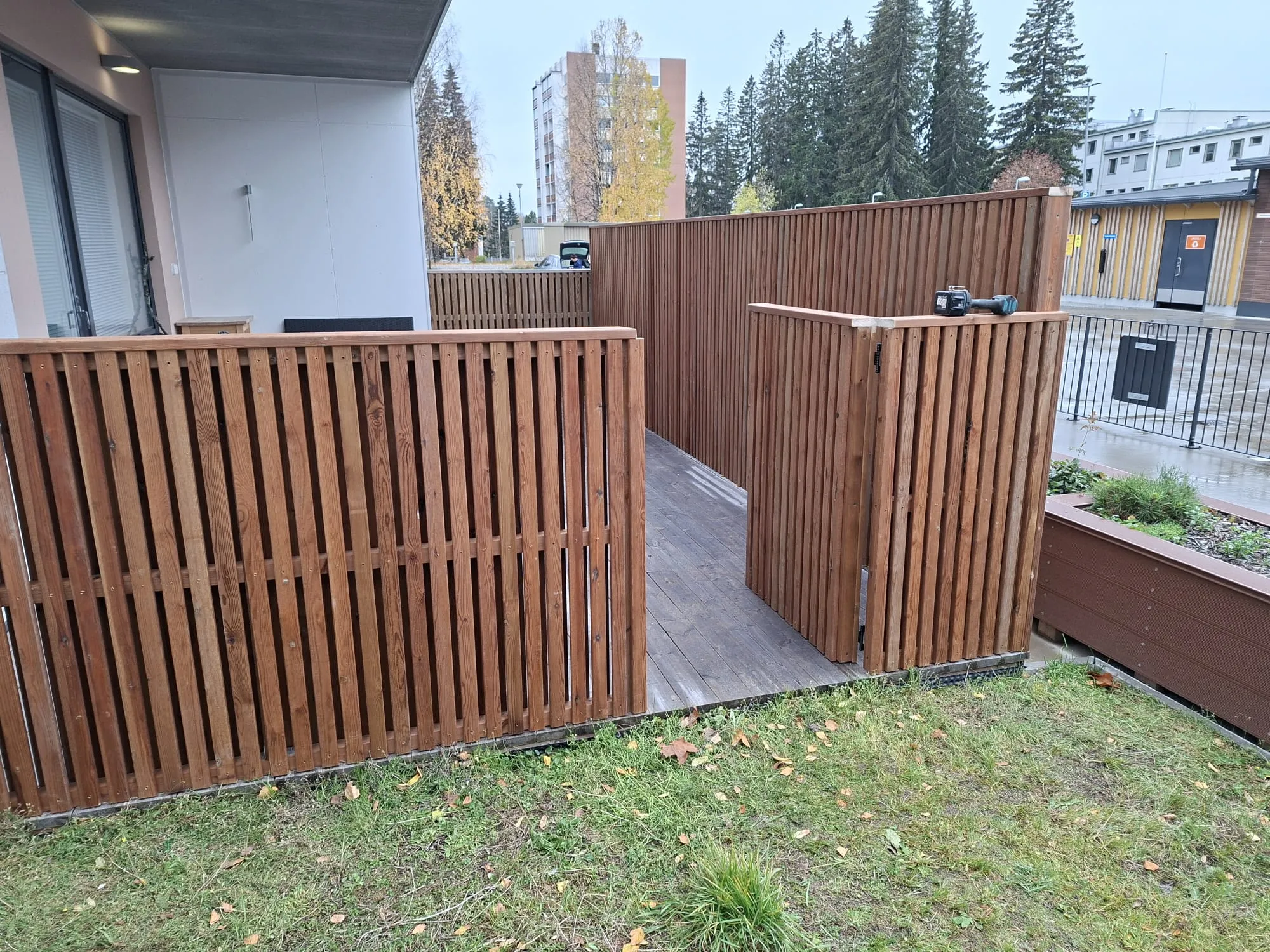 Ruskea pystysuorista laudoista rakennettu puuaita rajaa piha-aluetta, jossa on puinen terassi. Aidan ulkopuolella on vihreää nurmikkoa ja syksyisiä lehtiä. Taustalla näkyy kerrostalo, mäntyjä ja keltainen rakennus. Aidan päällä lepää työkalu. Kuva on otettu pilvisenä päivänä.