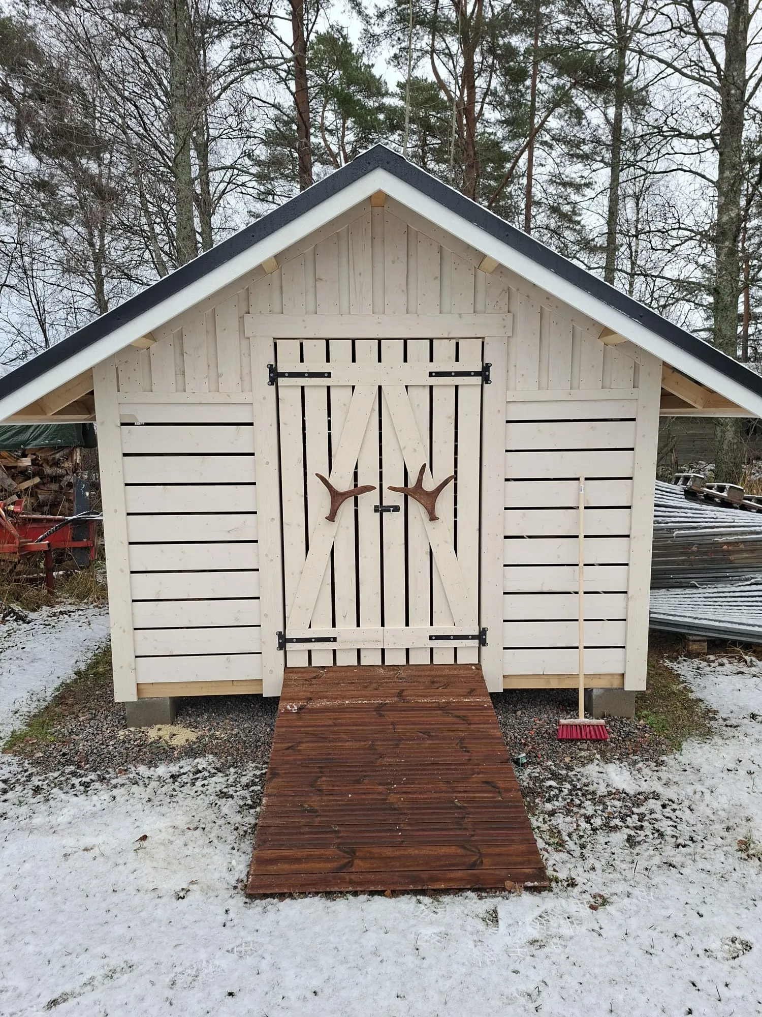 Vaalea puinen vaja tummalla katolla seisoo lumisessa maisemassa. Vajan pariovissa on mustat saranat ja sarvenmuotoiset kahvat. Oven edessä on tummanruskea puinen luiska. Maassa on laikukasta lunta ja soraa. Oikealla vajan seinää vasten nojaa punainen luuta. Taustalla näkyy paljaita lehtipuita ja havupuita pilvisenä päivänä.