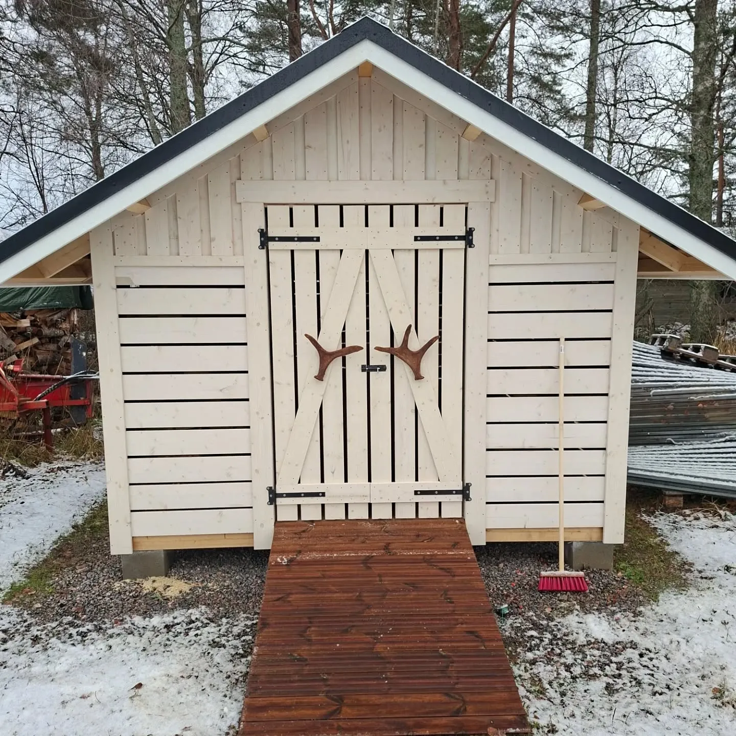 Vaalea puinen vaja tummalla katolla seisoo lumisessa, soraisessa maastossa. Vajan etuosassa on pariovet, joissa on sarvenmuotoiset kahvat. Oven edessä on tumma puinen lankkuramppi. Oikealla puolella nojaa punainen harja. Taustalla on paljaita puita ja pinottuja tavaroita.