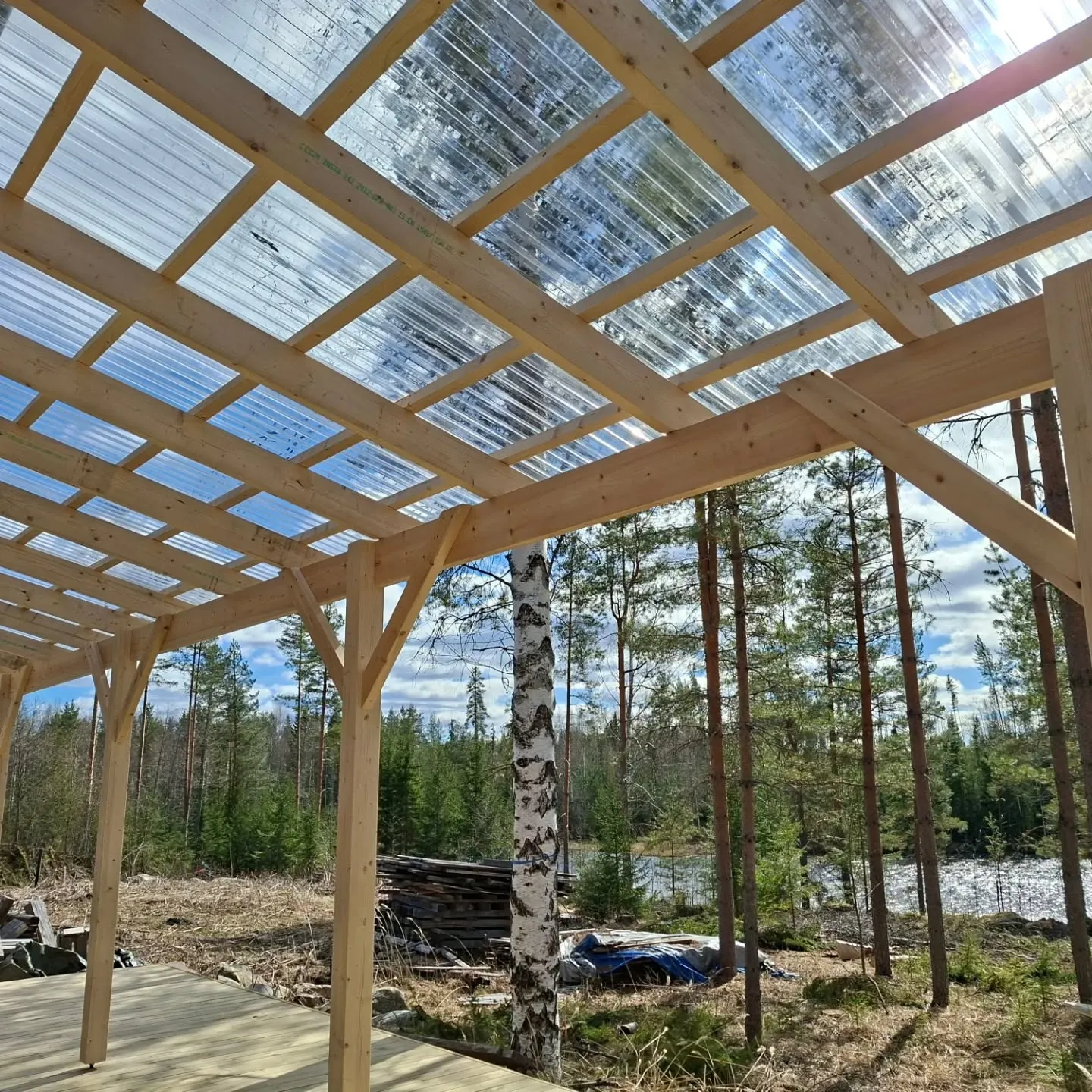 Vaaleasta puusta rakennettu terassin katos, jossa on läpinäkyvä aaltomuovikatto. Katon läpi näkyy sininen taivas ja valkoisia pilviä, ja kirkas auringonvalo heijastuu pinnasta. Katos avautuu metsäiseen maisemaan, jossa näkyy mäntyjä, kuusia ja koivun runko. Taustalla kimaltelee vesistö. Alhaalla on puinen terassilattia ja maassa kuivaa kasvillisuutta.