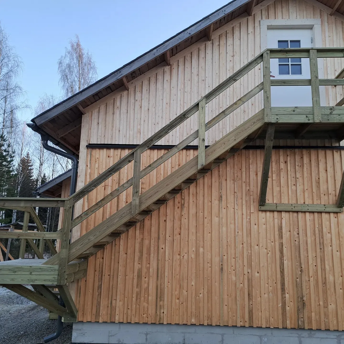 Vaaleanruskea puurakennus, jossa on tummanvihreät ulkoportaat ja kaiteet, jotka nousevat valkoiselle ovelle. Rakennuksen tumma katto ja musta vesikouru näkyvät ylhäällä. Alhaalla on harmaa betoninen perustus. Taustalla on paljaita lehtipuita ja havupuita vaaleansinistä taivasta vasten kirkkaassa päivänvalossa.