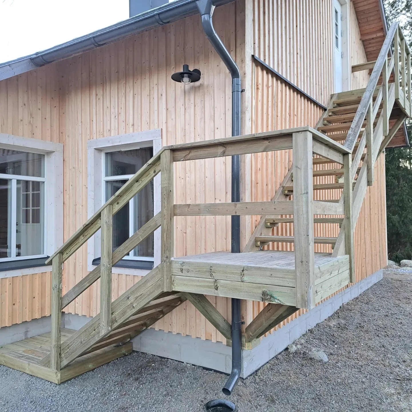 Vaaleapuinen pystylaudoitettu talon seinä, jossa on kaksi valkoista ikkunaa. Seinän edessä on vaaleanvihreästä kestopuusta rakennetut portaat ja tasanne, jotka nousevat yläkerran valkoiselle ovelle. Tummanharmaa syöksytorvi kulkee seinää pitkin portaiden takana. Seinällä on musta ulkovalaisin. Maassa on soraa ja talon perustukset ovat betoniharkkoja.