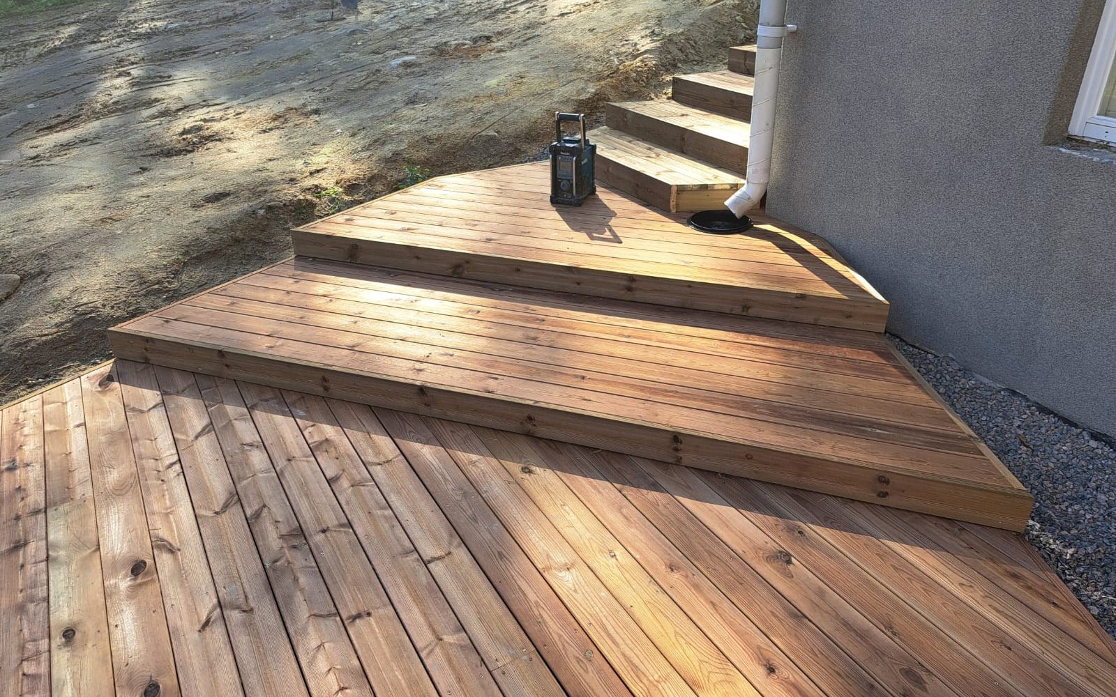 Aurinkoinen kuva vaaleanruskeasta puisesta terassista, jossa on useita askelmia. Terassilla on tumma lyhty. Oikealla on harmaa rakennuksen seinä valkoisella syöksytorvella ja soraa maassa. Vasemmalla näkyy hiekkainen rinne.
