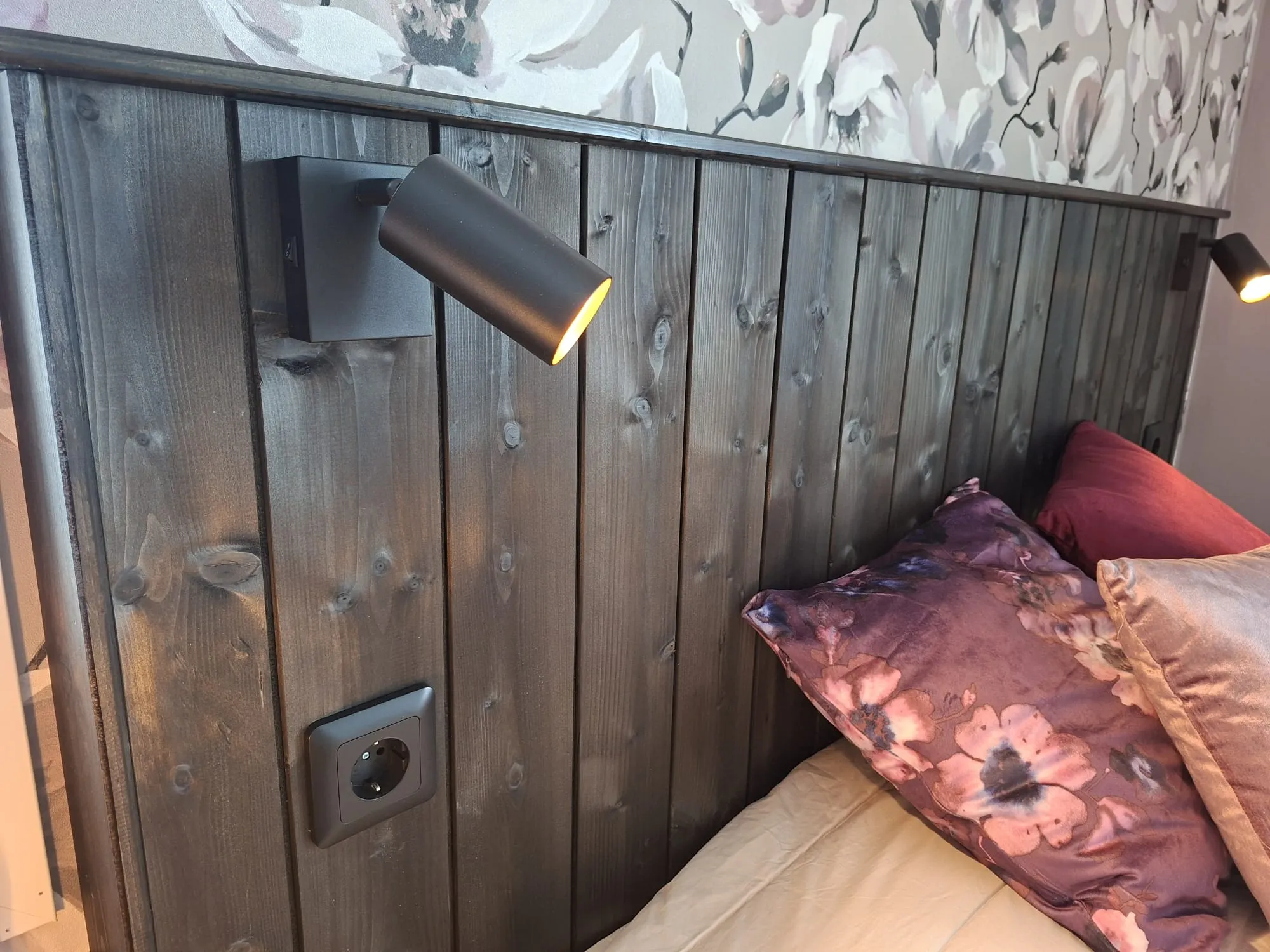 Kuva makuuhuoneen sängynpäädystä, joka on tehty tummista pystysuuntaisista puulaudoista. Vasemmalla on musta, päällä oleva lukuvalo, joka hehkuu lämmintä valoa, ja sen alapuolella musta pistorasia. Päädyn yläpuolella seinällä on tapetti, jossa on suuria harmaita, valkoisia ja vaaleanpunaisia kukkakuvioita vaalealla pohjalla. Sängyllä on useita tyynyjä, mukaan lukien violetti kukkakuvioinen tyyny etualalla ja vaaleanpunainen samettityyny oikealla.