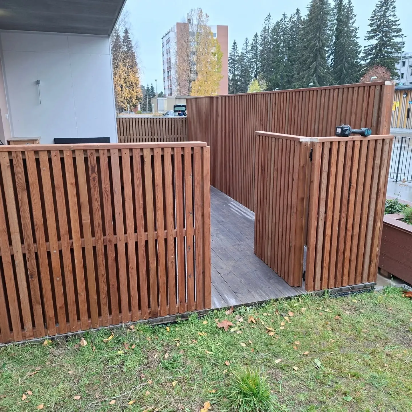 Ruskea puuaita ympäröi ulkoaluetta, jossa on tumma puulattia. Aita koostuu pystysuorista laudoista ja siinä on avoin portti oikealla. Etualalla on vihreää nurmikkoa syksyn lehdillä. Taustalla näkyy valkoinen rakennus, puita ja kerrostaloja. Oikeanpuoleisen aidan päällä on akkuporakone. Valo on tasaista päivänvaloa.