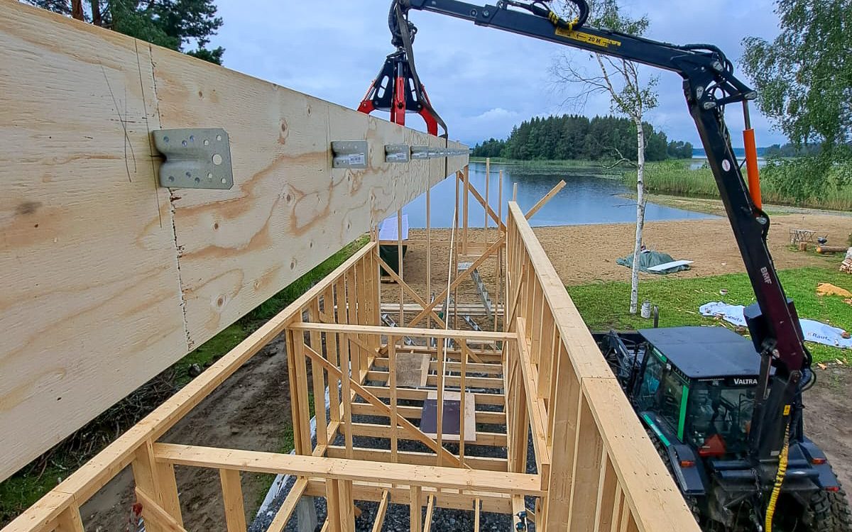 Kuva näyttää rakennustyömaan, jossa musta nosturivarsi, jossa on punainen tarttuja, nostaa suurta vanerista puupalkkia. Palkki on laskeutumassa osittain valmiin puurungon päälle. Oikealla alhaalla näkyy vihreä Valtra-traktori, johon nosturi on kiinnitetty. Taustalla on järvi, hiekkaranta ja metsäinen ranta pilvisenä päivänä.