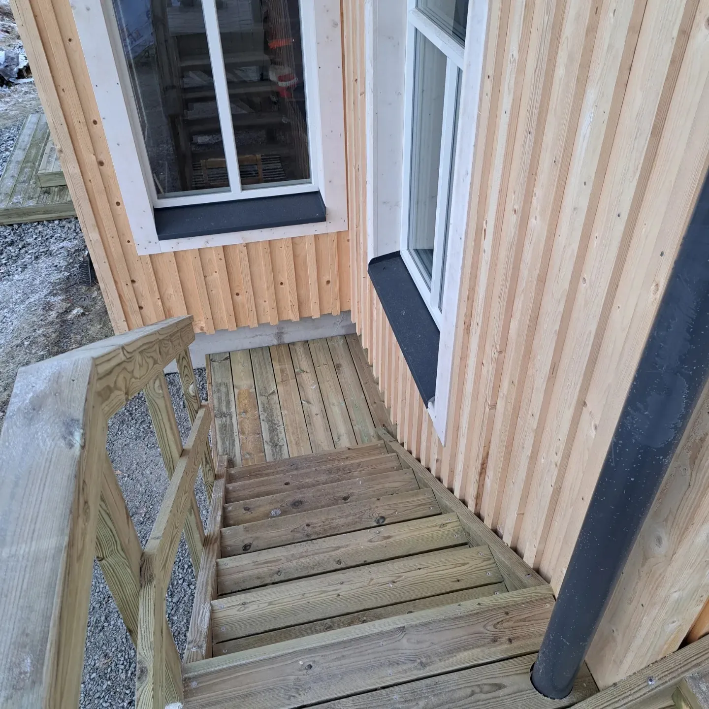 Yläviistosta otettu kuva puisista portaista, jotka laskeutuvat alas vaaleasta pystypaneloidusta puurakennuksesta. Rakennuksessa on valkoiset ikkunankarmit ja mustat ikkunalaudat. Portaissa on vihertävän harmaa puukaide vasemmalla ja oikealla näkyy musta syöksytorvi. Maassa on soraa ja lunta.