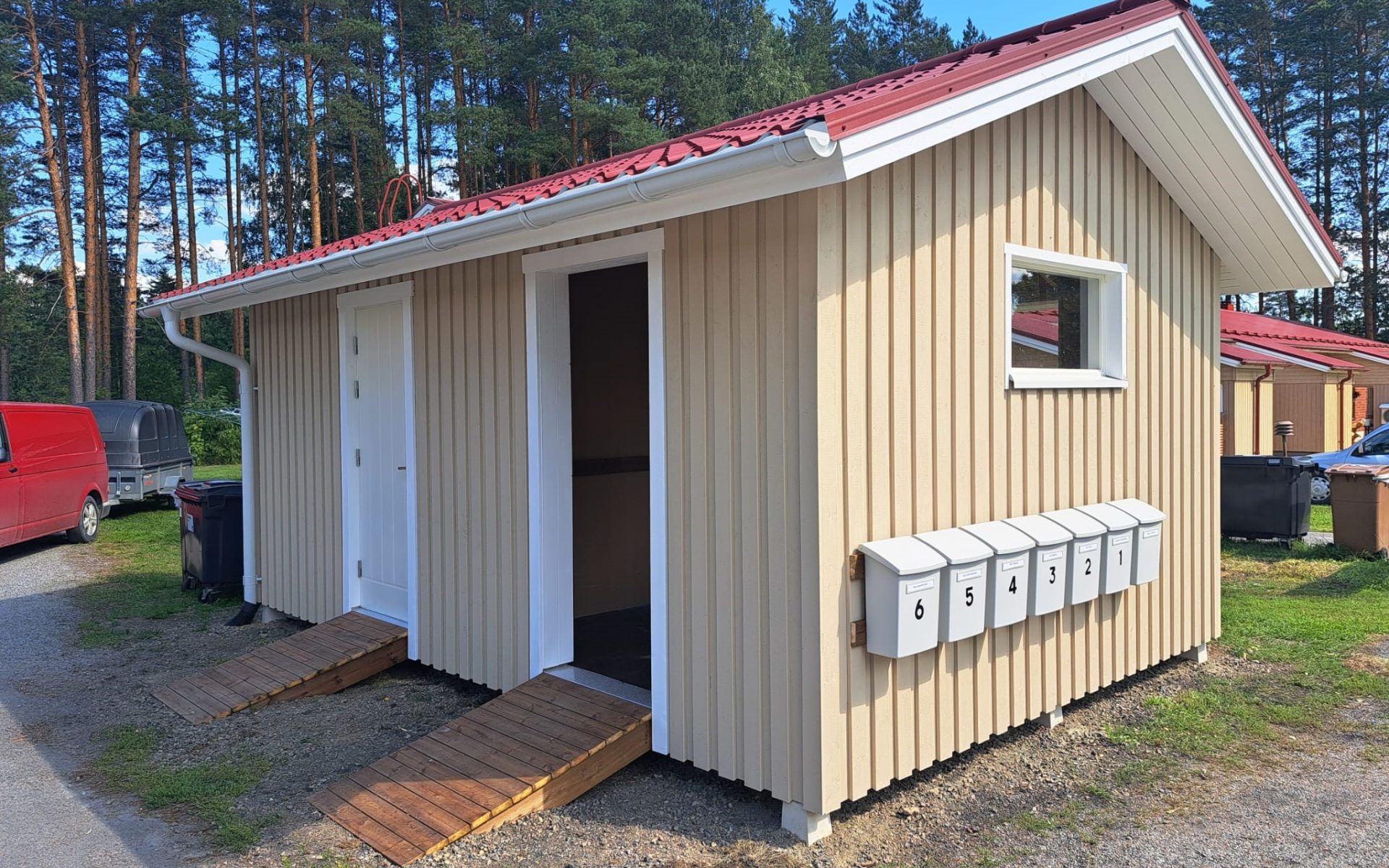 Vaaleankeltainen puurakennus punaisella peltikatolla ja valkoisilla ränneillä. Rakennuksessa on kaksi valkoista ovea, joihin johtaa puiset luiskat. Oikealla seinällä on pieni ikkuna ja kuusi numeroitua valkoista postilaatikkoa. Taustalla on metsää ja sininen taivas. Vasemmalla näkyy punainen pakettiauto ja musta peräkärry. Aurinkoinen sää.
