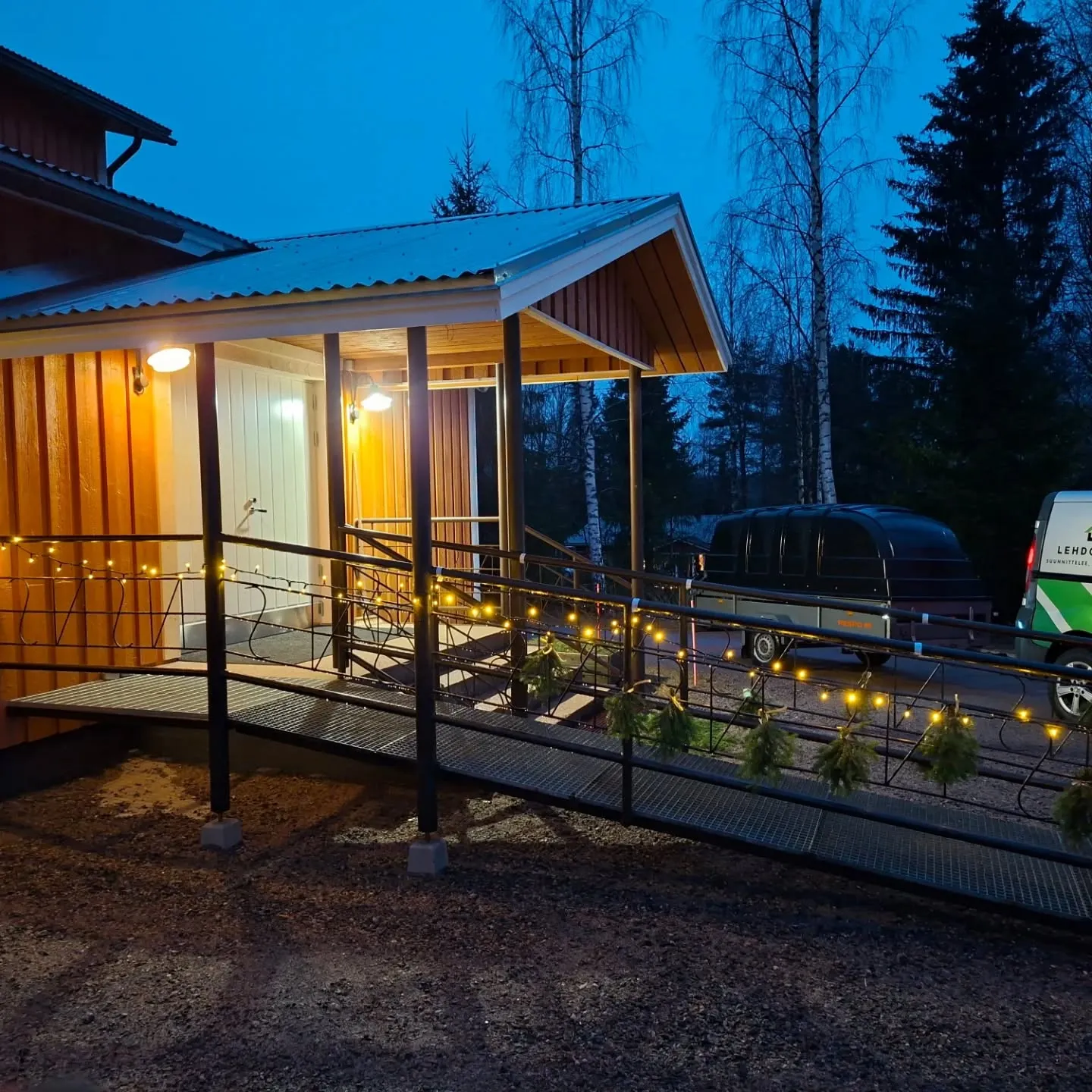 Iltakuva puurakennuksen sisäänkäynnistä. Rakennuksessa on oranssinruskeat seinät, vaalea ovi ja aaltopeltikatto. Kuistin yläpuolella on kaksi lämmintä keltaista valaisinta. Sisäänkäynnille johtaa metallinen ramppi mustalla kaiteella, jonka varrella on rivi pieniä keltaisia valoja ja pieniä kuusenoksia. Taustalla näkyy puita ja tumma iltataivas. Oikealla on tumma peräkärry ja osittain valkoinen pakettiauto.
