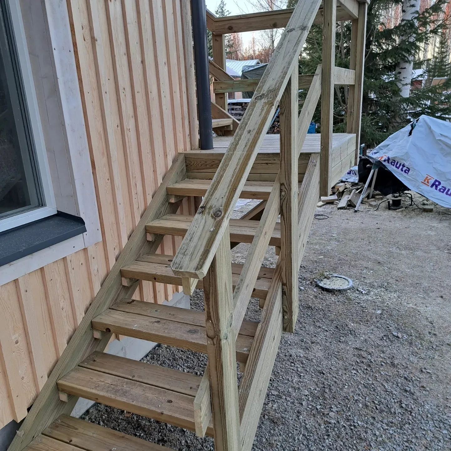 Vaalean puutalon pystypaneeliseinän vieressä on puusta rakennetut portaat ja kaiteet, jotka johtavat ylös terassille. Vasemmalla näkyy ikkuna tummalla ikkunalaudalla. Maassa on harmaata soraa, jossa on hieman kuuraa. Taustalla näkyy puita ja valkoinen pressu rakennustarvikkeiden päällä. Kuvan valaistus on luonnollinen ja pehmeä.
