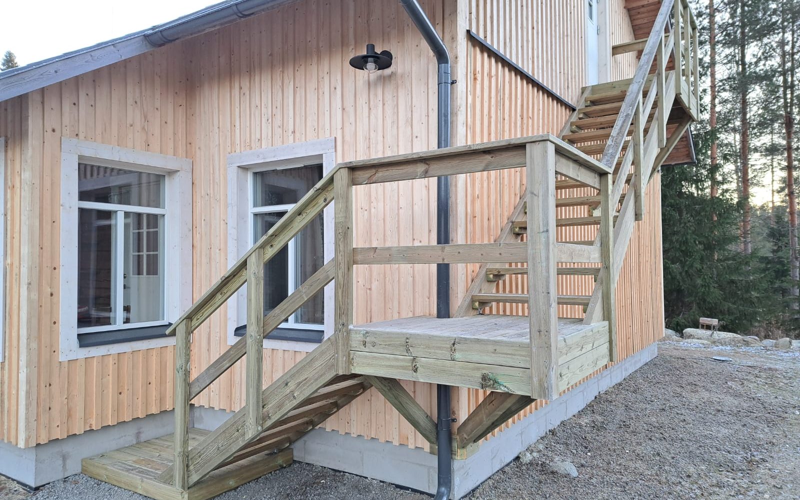 Vaaleapuinen talo pystypaneloinnilla ja valkoisilla ikkunankarmeilla. Talon edessä on vihertävänruskeat puiset ulkoportaat, jotka nousevat yläkertaan. Seinällä näkyy harmaa syöksytorvi ja musta ulkovalo. Maassa on soraa ja taustalla mäntymetsää ilta-auringossa.