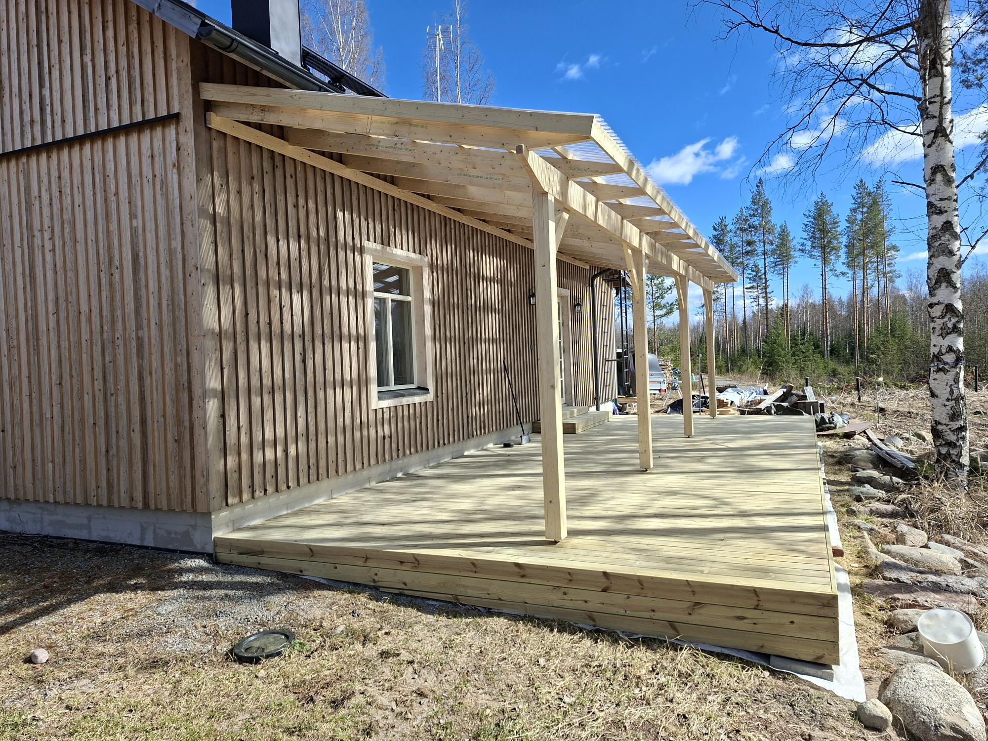 Aurinkoinen kuva puutalosta, jossa on vaaleanruskea pystylautaverhous ja uusi puinen terassi. Terassilla on läpinäkyvä katos, jota tukevat vaaleat puupylväät. Taustalla näkyy metsää ja oikealla on koivu sinistä, pilvistä taivasta vasten.