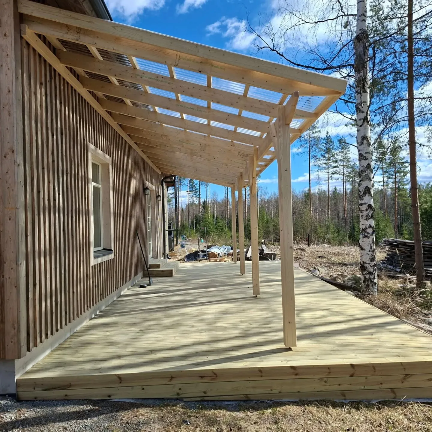 Uusi puutalo, jossa on vaaleasta puusta rakennettu suuri terassi ja läpinäkyvällä katolla varustettu pergola. Aurinkoinen päivä, sininen taivas ja metsäinen tausta, jossa näkyy koivu ja mäntyjä.