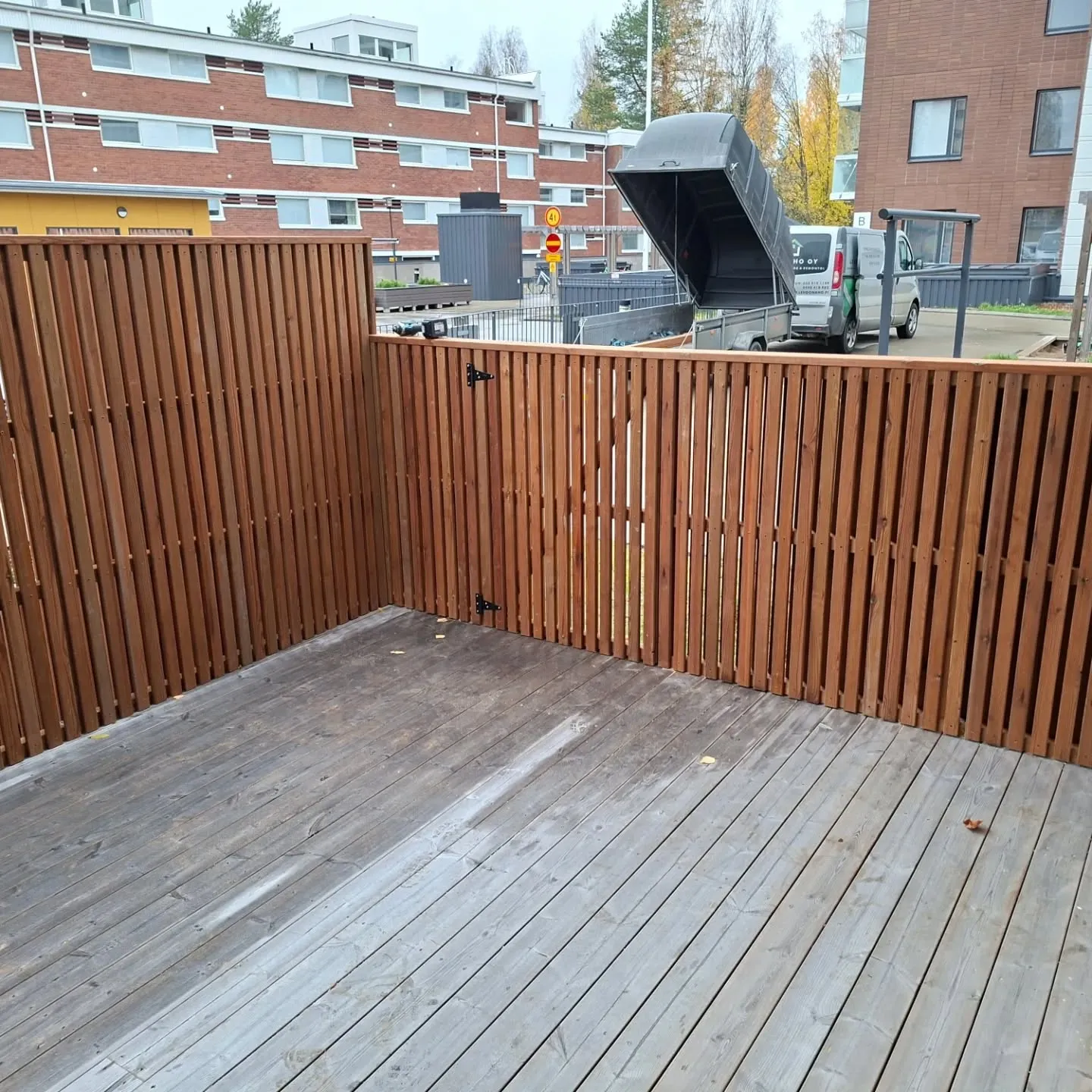 Kuvassa on ulkoterassi, jonka harmaat puulaudat ovat kuluneet. Terassia rajaa L-muotoinen ruskea puuaita. Aitojen takana näkyy asuinalue, jossa on punatiilisiä kerrostaloja. Keskellä taustalla on valkoinen pakettiauto, johon on kytketty musta peräkärry, jonka takalaita on nostettu ylös. Päivä on pilvinen ja valaistus tasainen.
