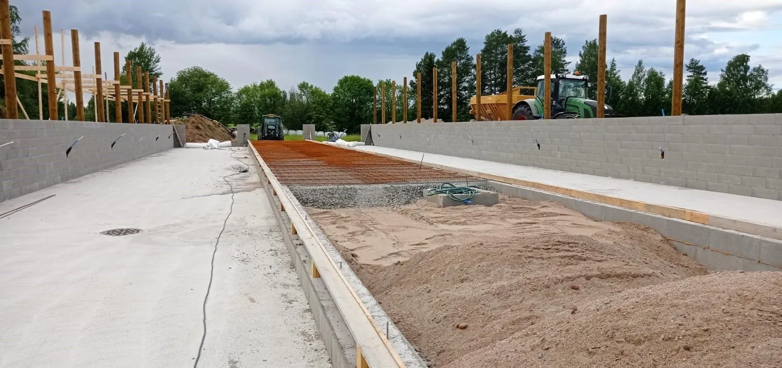 Rakennustyömaa, jossa on kaksi pitkää betoniharkkoseinää. Keskellä on hiekkaa, soraa ja raudoitusverkkoa valmiina valua varten. Vasemmalla on valettu betonilattia viemärillä. Seinien yläpuolella näkyy puurunkoja ja taustalla kaksi traktoria puiden ja pilvisen taivaan alla.