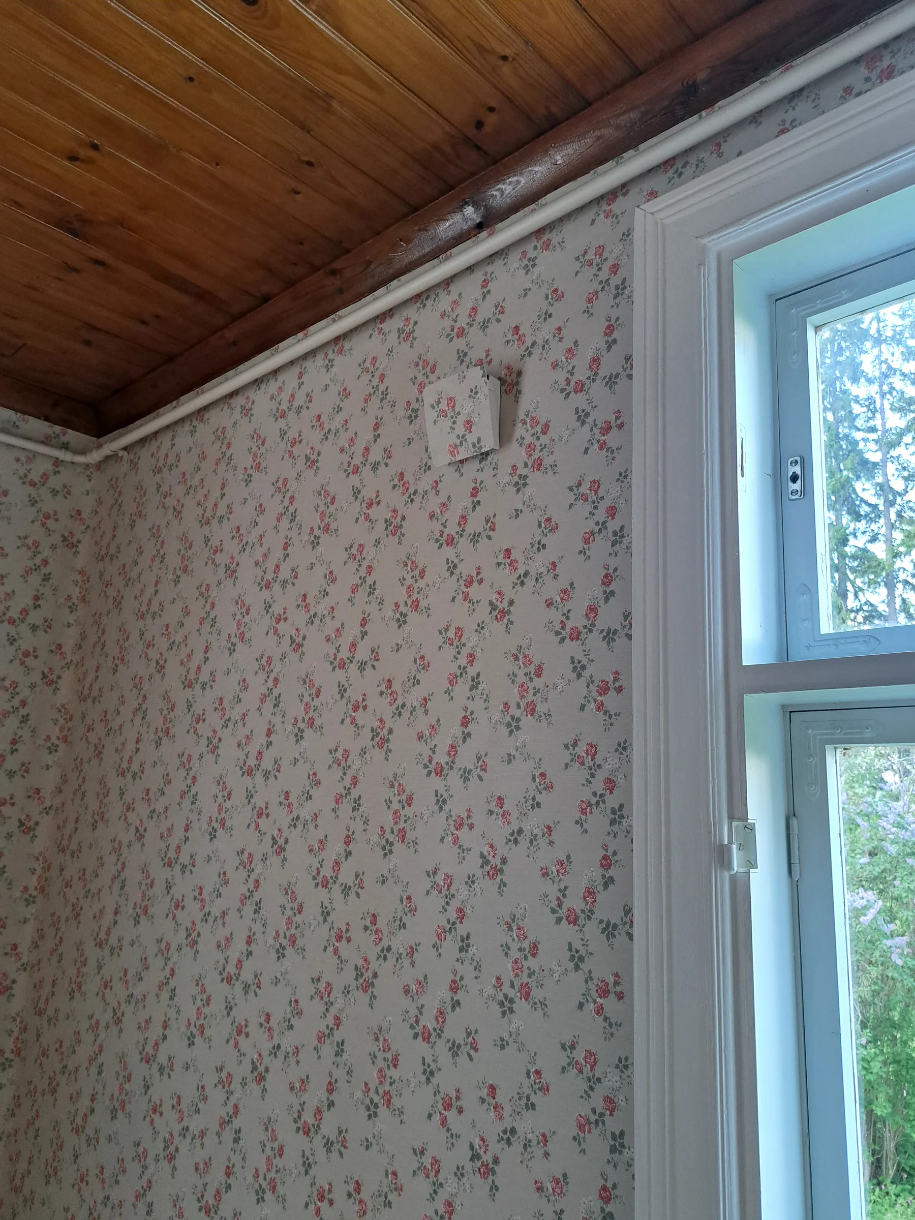 Huoneen sisäkuva, jossa on tummanruskea puukatto ja vaalea, ruusukuvioinen tapetti seinillä. Katon rajassa kulkee tumma puupalkki ja valkoinen putki. Seinällä on myös pieni, tapetilla päällystetty laatikko. Oikealla on valkoinen ikkuna, josta näkyy ulos vihreää puustoa ja pensaita.