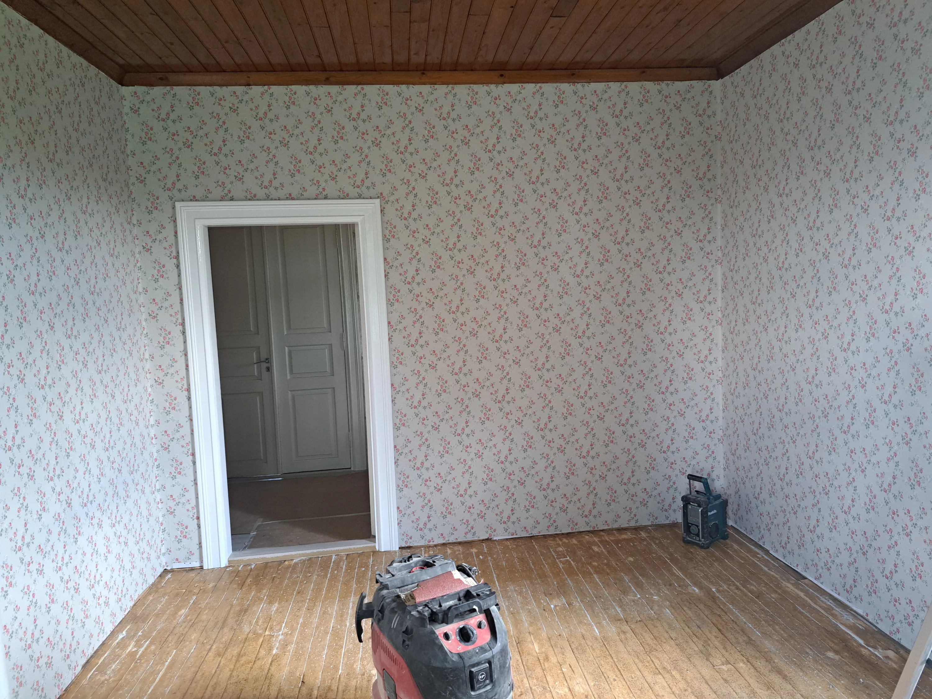 Huone, jossa on vaaleat seinät pienillä punaisilla ruusuilla ja vihreillä lehdillä. Katossa on tummanruskeat puupaneelit. Lattialla on vaaleat puulankut, joissa on valkoisia jälkiä. Oikealla etualalla on punamusta työkalu ja oikealla takana tumma radio. Valkoinen oviaukko johtaa toiseen huoneeseen, jossa näkyy harmaa ovi.