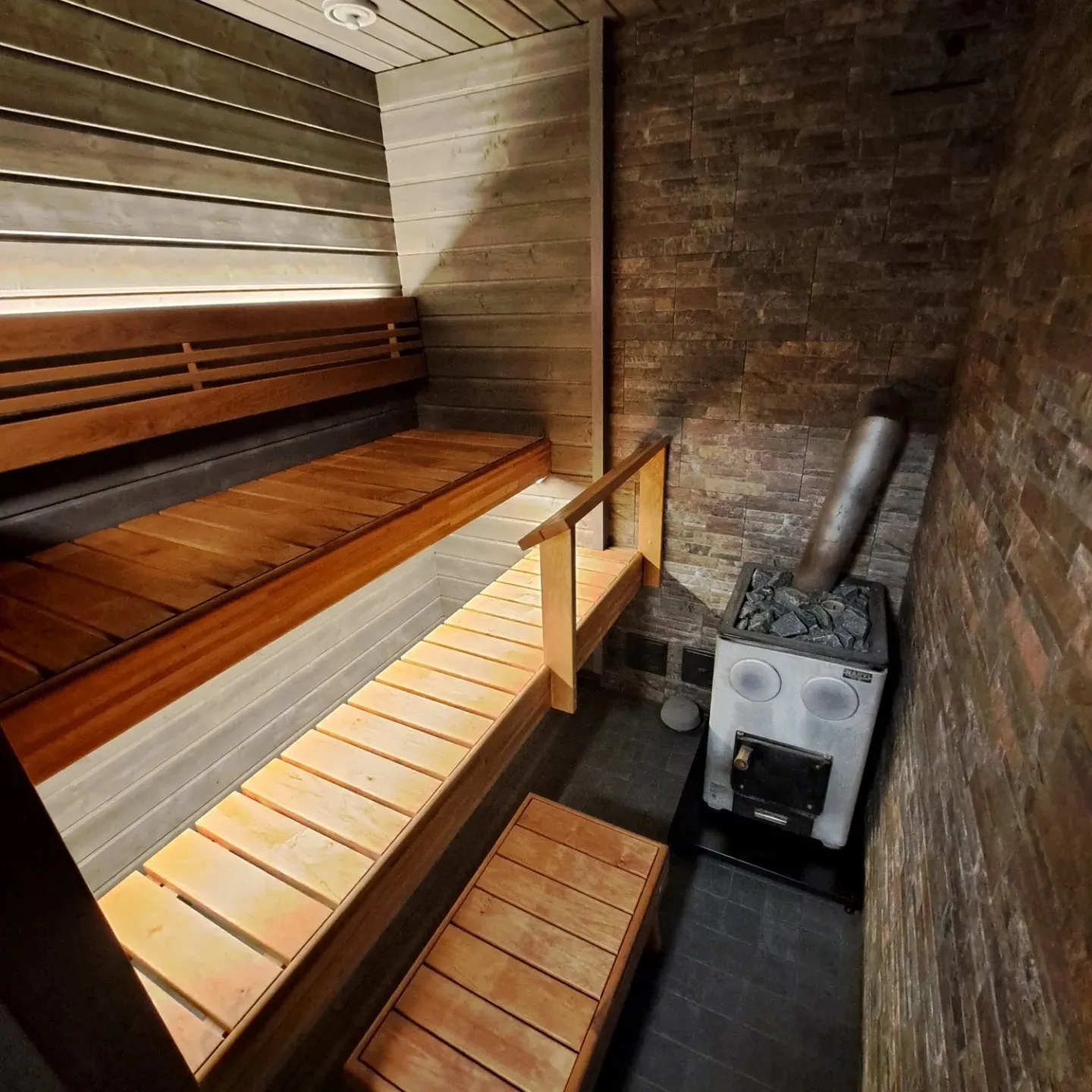 Moderni sauna, jossa on puuseinät ja kivilaattaseinä. Useita ruskeita puulauteita, joissa on lämmin epäsuora valaistus. Oikealla on harmaa puulämmitteinen Harvia-kiuas kiuaskivineen ja metallinen savupiippu. Lattia on tummaa laattaa.
