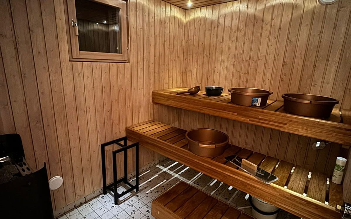 Saunan sisätila, jossa vaaleat puuseinät ja kaksi tasoa puisia lauteita. Vasemmalla seinällä on pieni ikkuna. Ylemmällä lauteella on puinen kauha, tumma kulho ja kaksi kuparinväristä pesuvatia. Keskimmäisellä lauteella on yksi kuparinvärinen pesuvati, metallitarjotin harjalla ja valkoinen pullo. Vasemmalla alhaalla on musta kiuas ja oikealla musta metalliteline. Lattia on valkoista laattaa. Tilaa valaisee lämmin, epäsuora valaistus ylhäältä ja lauteiden alta.