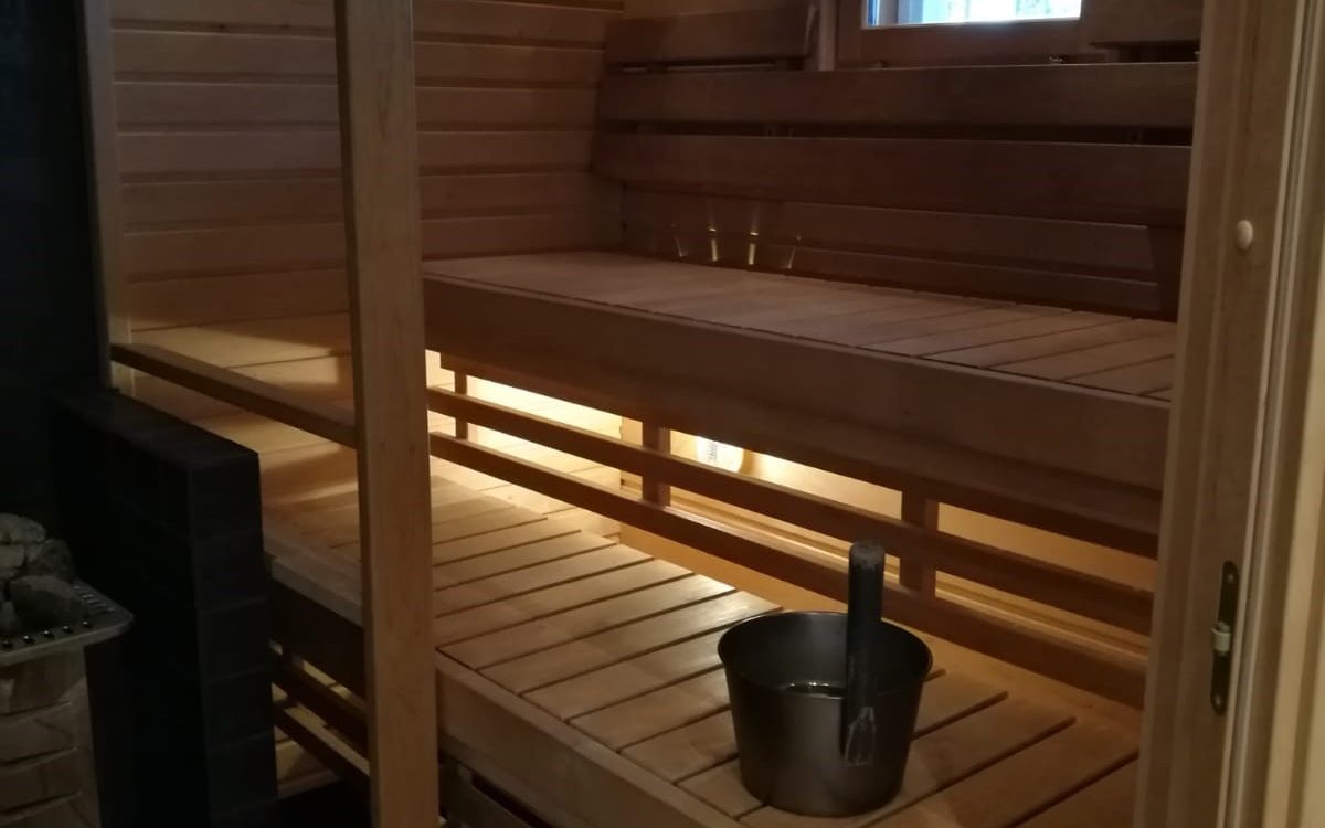 Saunan sisätila, jossa on vaaleat puupenkit kahdessa tasossa ja puuseinät. Vasemmalla näkyy kiuas kivineen. Lattialla on metallinen löylyämpäri ja kauha. Lämmin, epäsuora valaistus penkkien alta ja ikkuna ylhäällä valaisevat tilaa.
