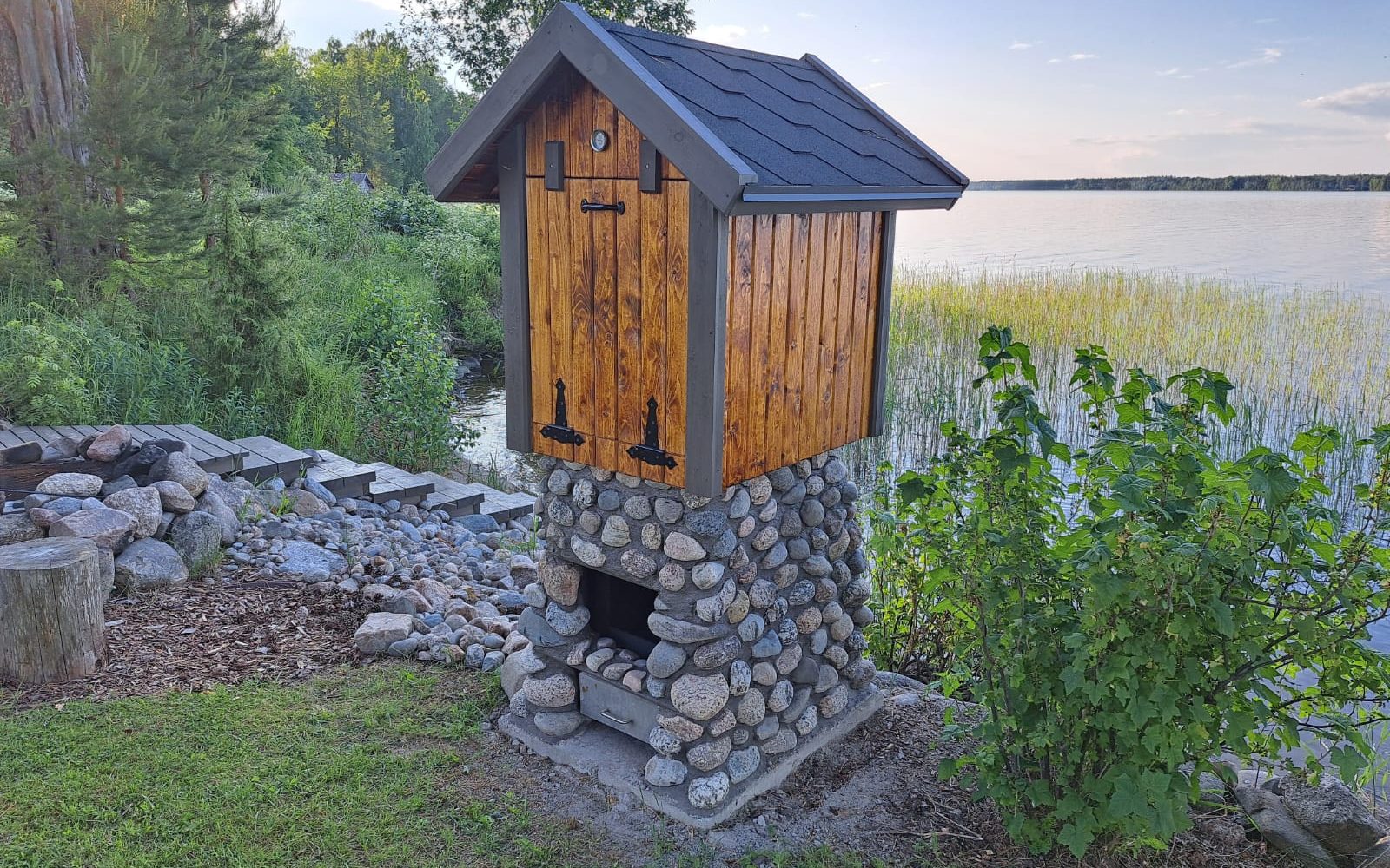 Puinen savustamo tummalla katolla seisoo kivijalustalla järven rannalla. Savustamon etuosassa on lämpömittari ja kivijalustan alaosassa on pieni luukku ulosvedettävällä laatikolla. Ympärillä on vihreää kasvillisuutta, puita, kiviä ja puiset portaat, jotka johtavat kohti vettä. Taustalla avautuu laaja järvimaisema kaislikkoineen ja kaukaisine metsänreunoineen. Kuva on otettu kirkkaassa, aurinkoisessa luonnonvalossa.