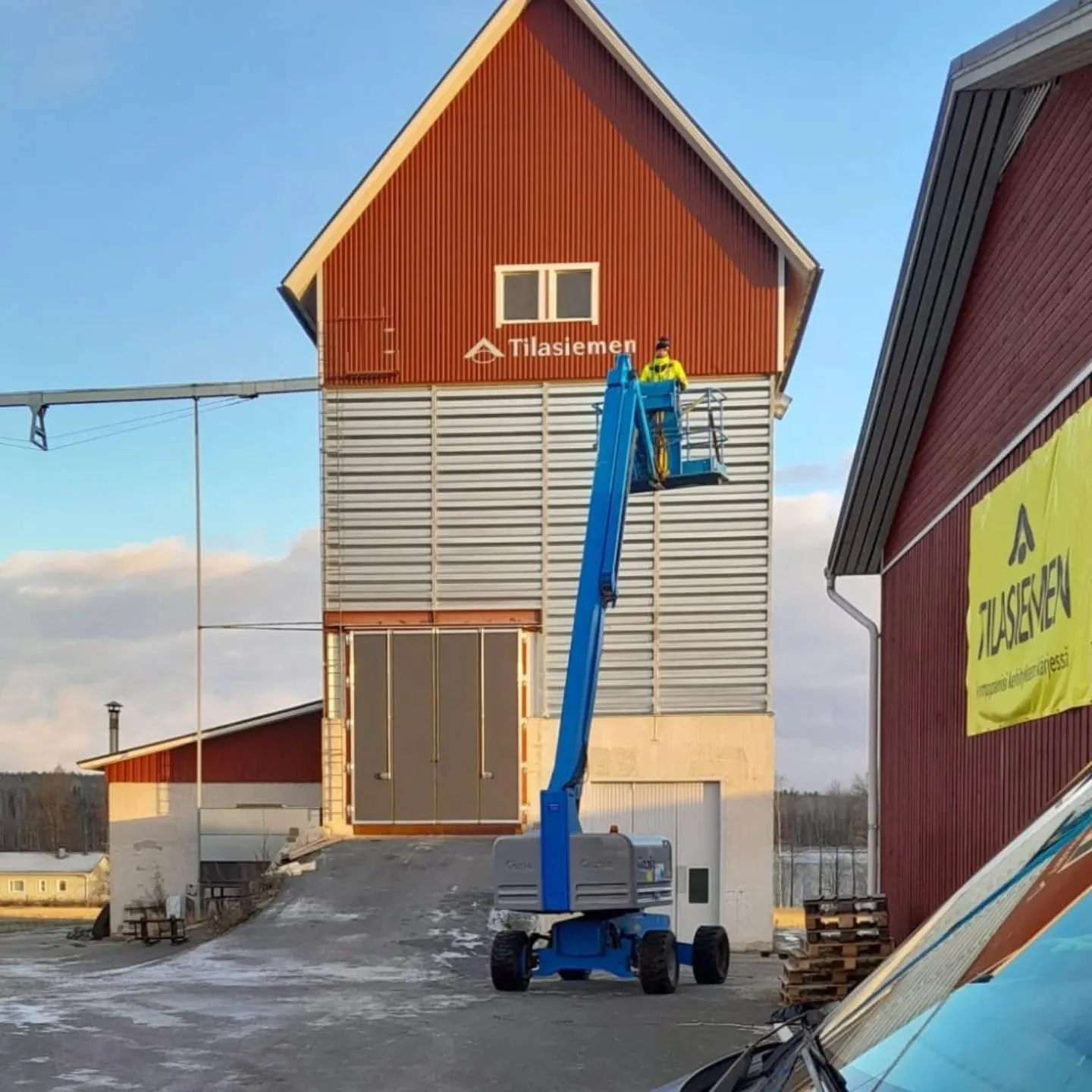 Päiväkuva korkeasta punaharmaasta rakennuksesta, jonka edessä sinisellä henkilönostimella työskentelee keltainen takki päällä oleva henkilö. Rakennuksen yläosassa lukee 'Tilasiemen'. Oikealla on toinen punainen rakennus, jossa on keltainen 'Tilasiemen'-banneri. Maassa on lumilaikkuja ja taivas on kirkas.